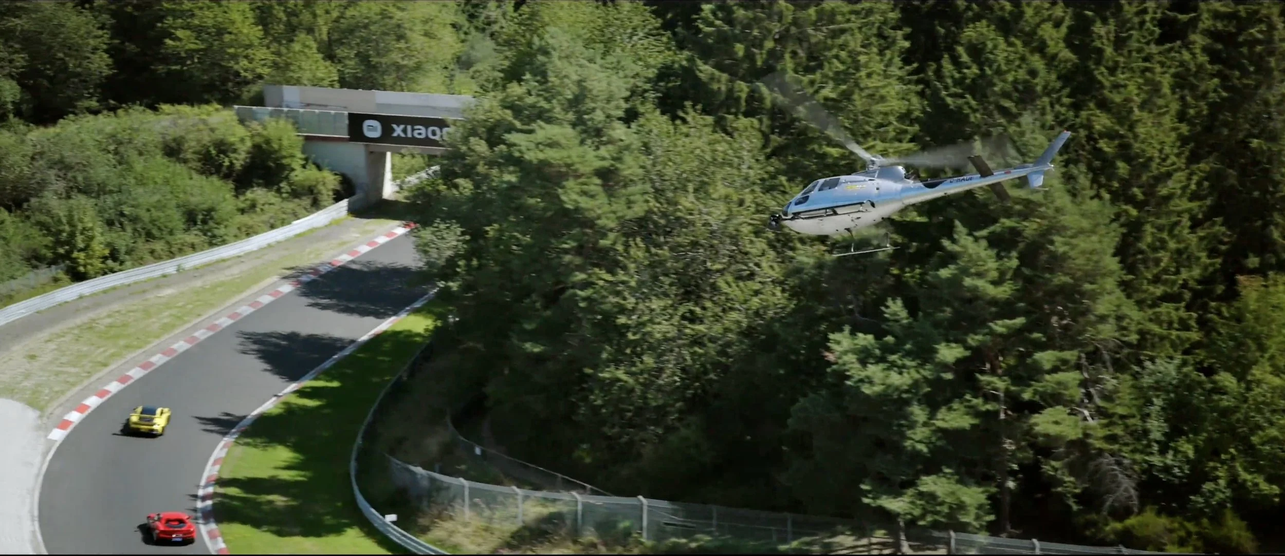 A helicopter flying near a race track with cars, surrounded by green trees.