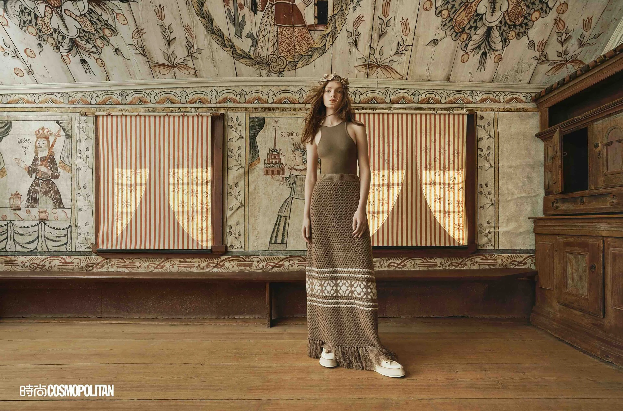 A woman standing in a room with ornate, historic-looking wall murals and wooden furniture, wearing a sleeveless earthy-toned top, a long patterned skirt, and white sneakers.