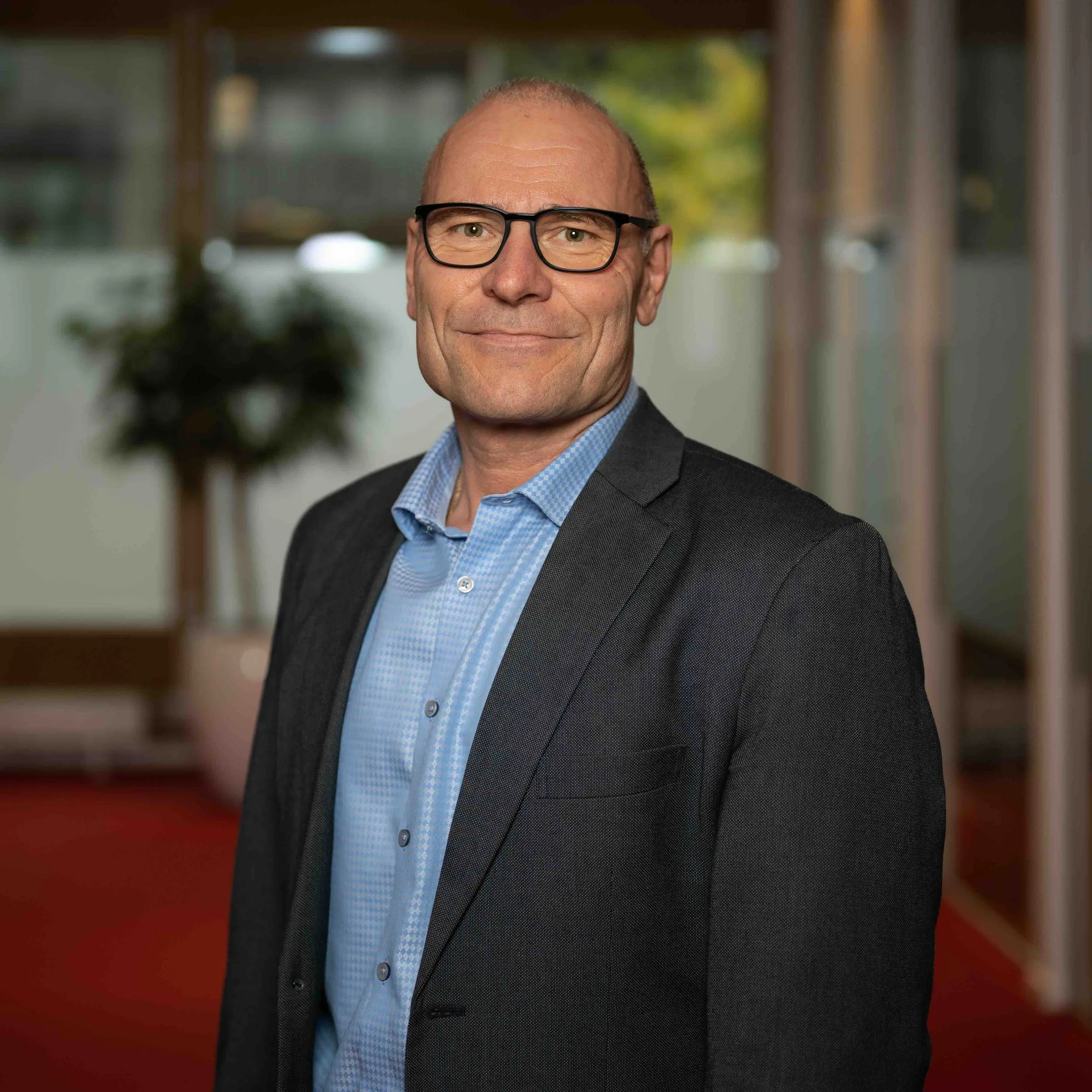 A middle-aged man with glasses and a tidily groomed appearance, wearing a dark suit jacket and a blue collared shirt, standing in an office setting with a blurred background and a potted plant.