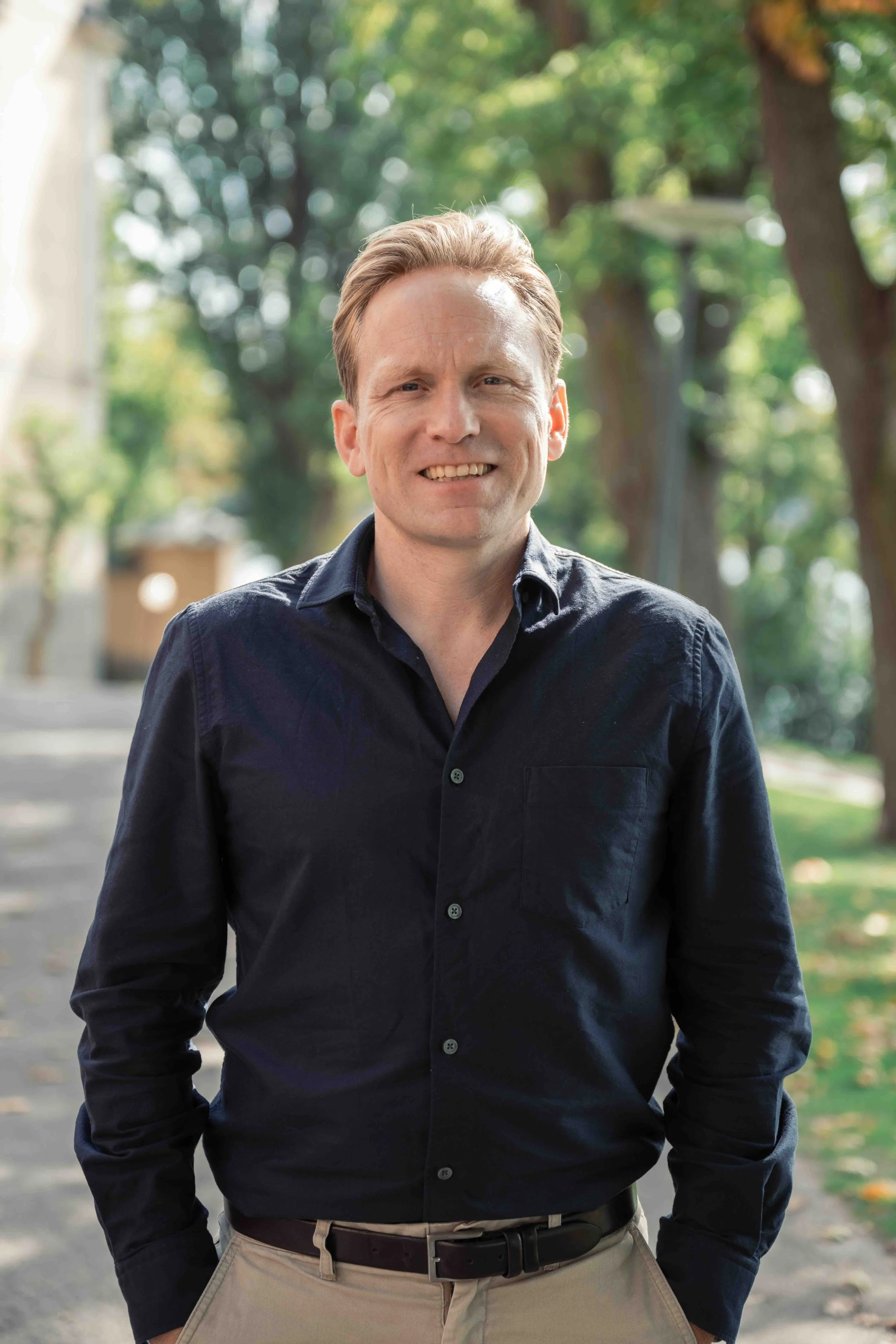 A man with short blond hair wearing a dark blue button-up shirt and beige pants stands outdoors in a park with trees and a pathway in the background.