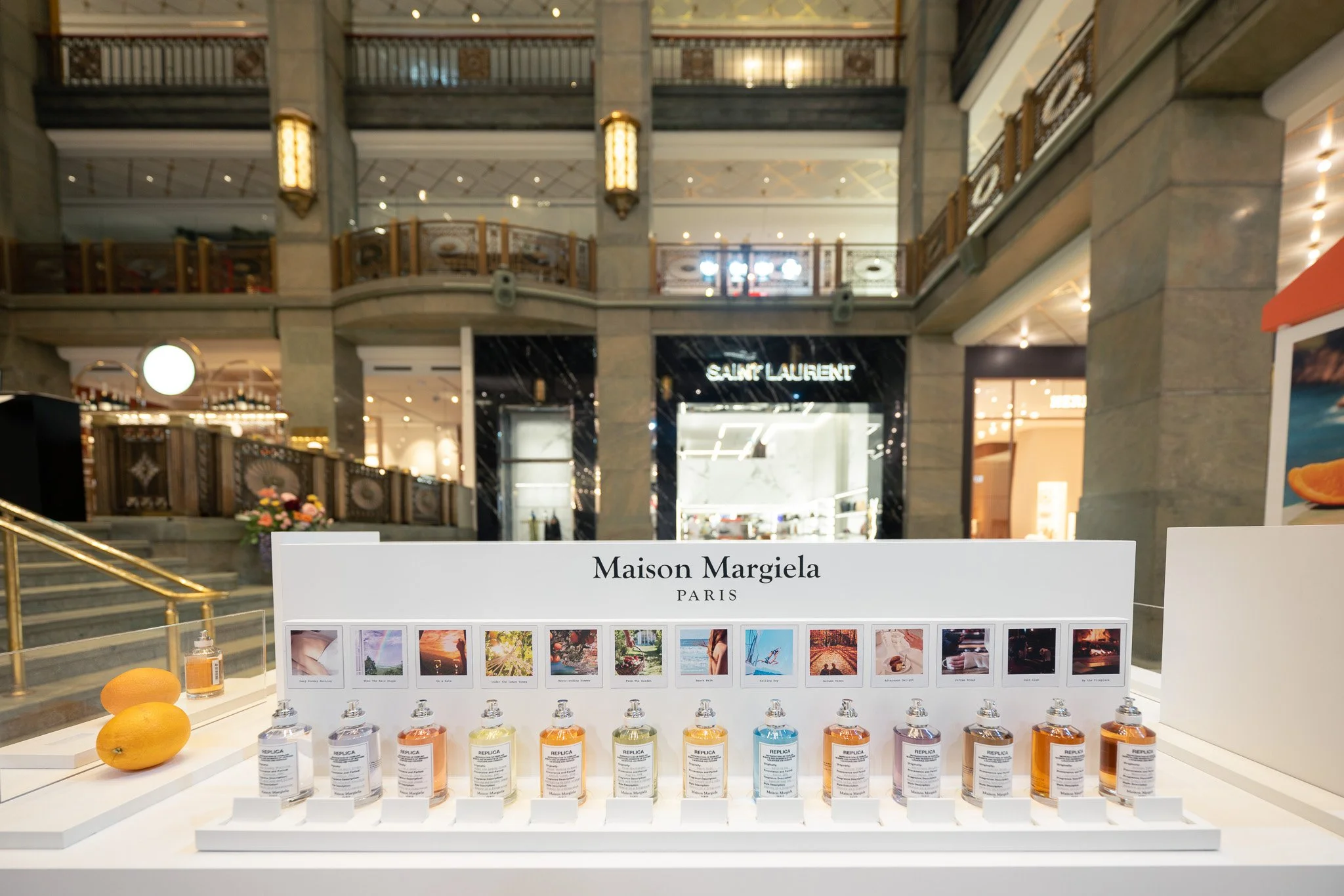 Display of Maison Margiela perfumes with colorful bottles arranged in a row in a shopping mall, with the store Saint Laurent visible in the background.
