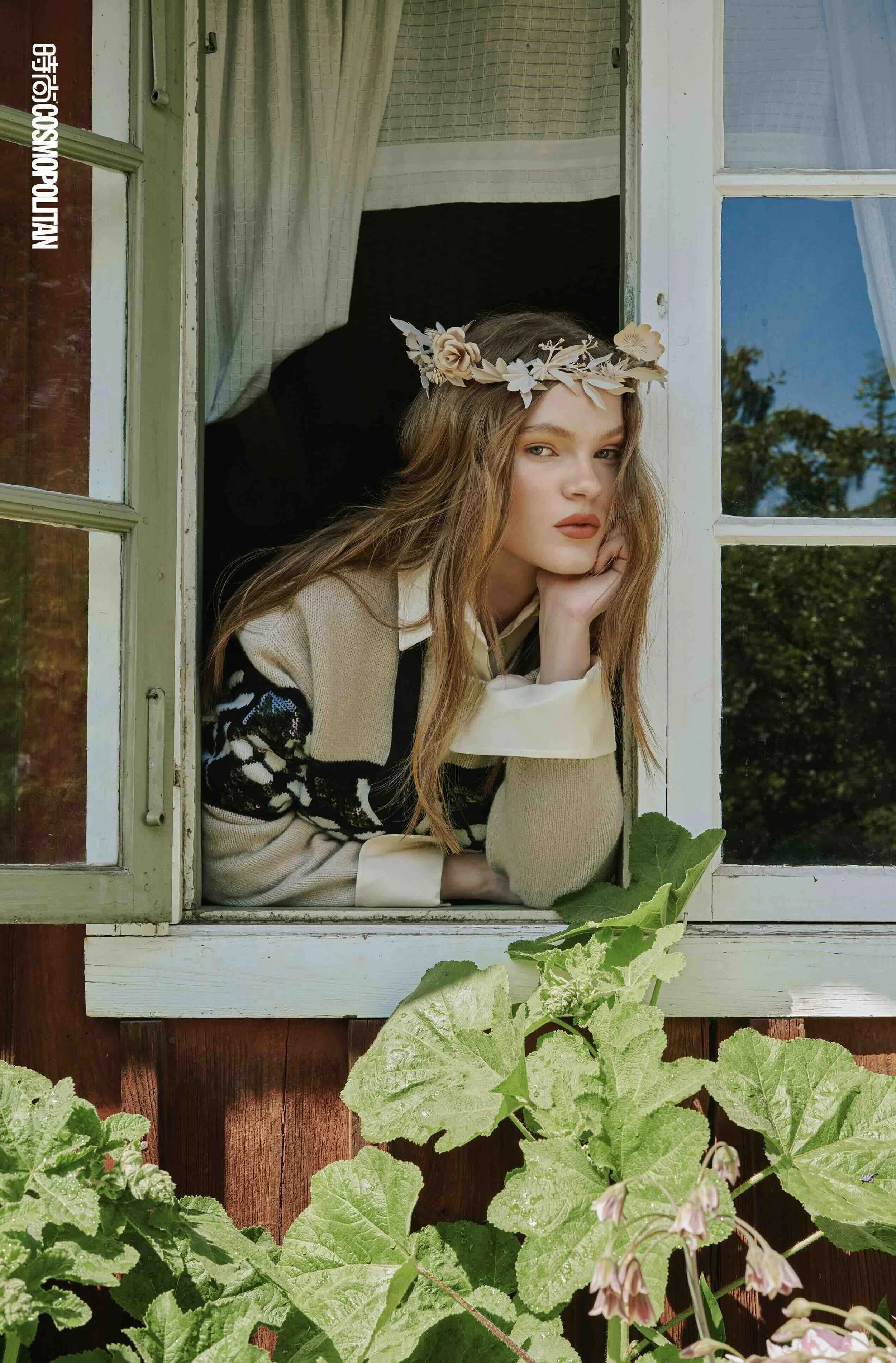A young woman with long wavy hair wearing a flower crown and patterned sweater leans on her hand, gazing out of a window of a rustic house in daylight, with green plants outside.