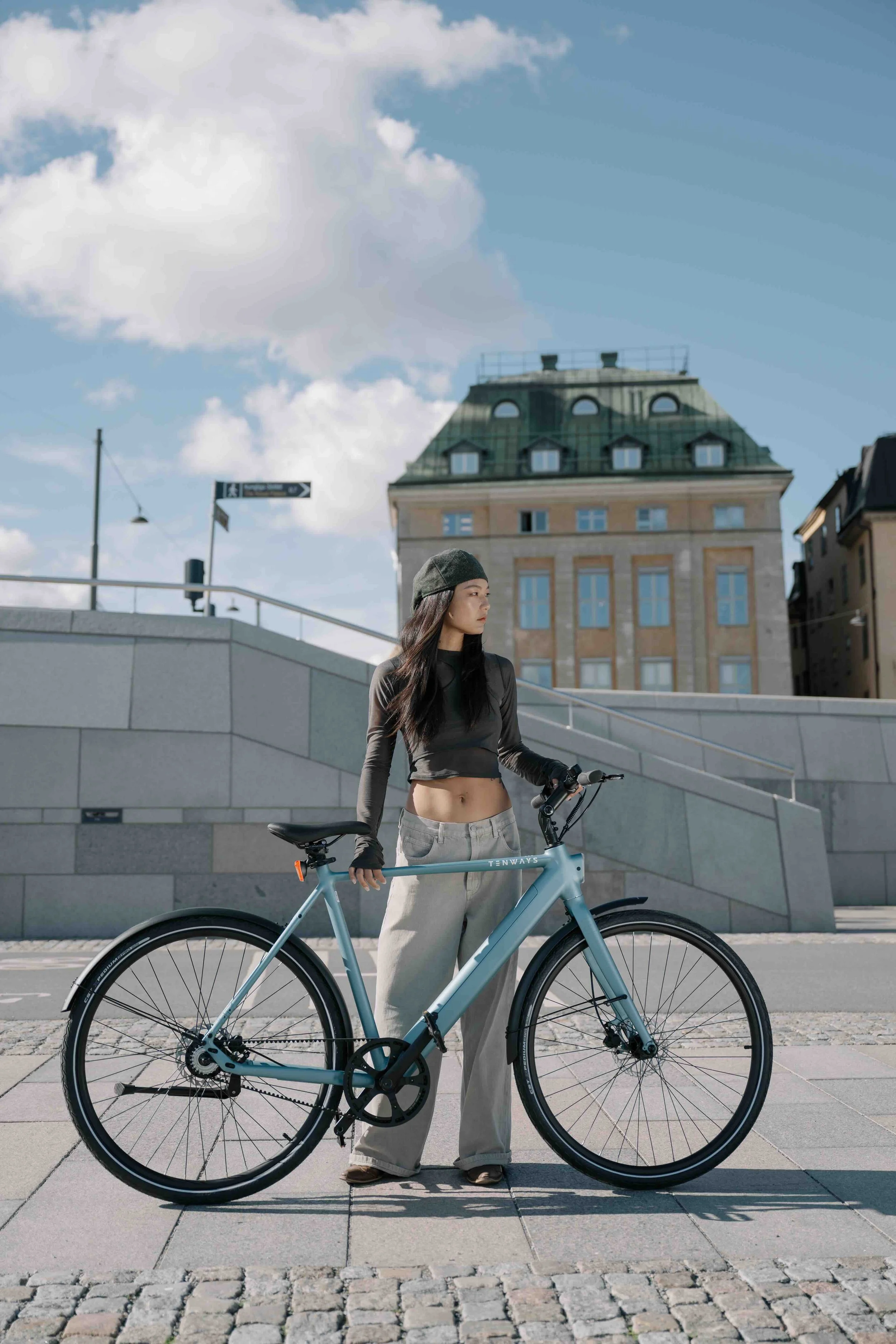 A young woman with long dark hair, wearing a gray beanie, long sleeve black crop top, beige pants, and black gloves, standing outdoors on a paved sidewalk, holding a blue bicycle with black tires and gears, with a building and cloudy sky in the background.
