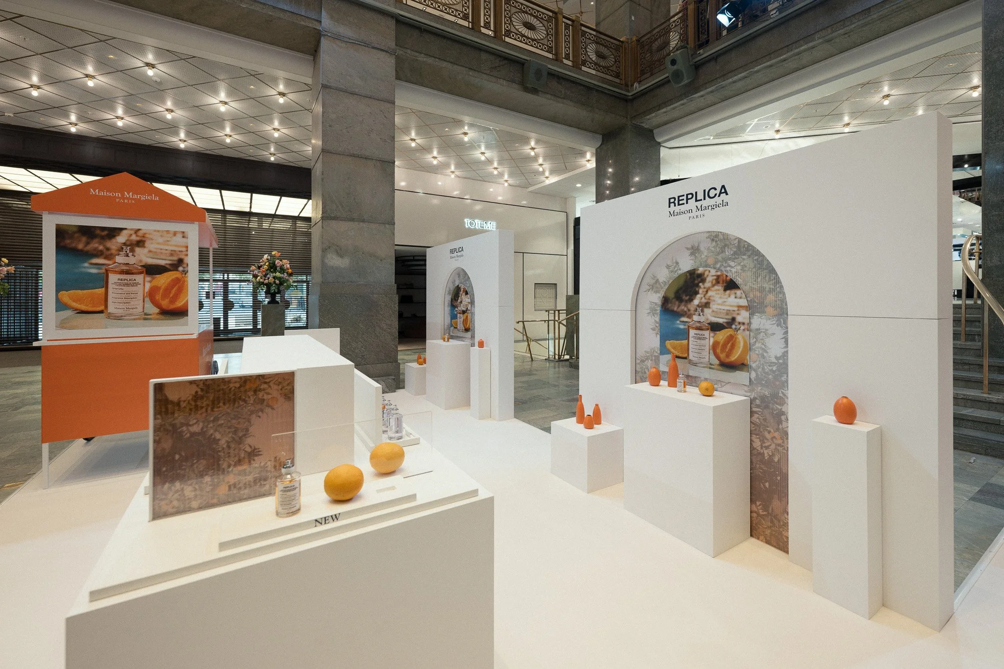 Exhibition display for Maison Margiela's Replica fragrance, featuring minimalist white backdrop, decorative oranges and bottles, with branding and promotional images.