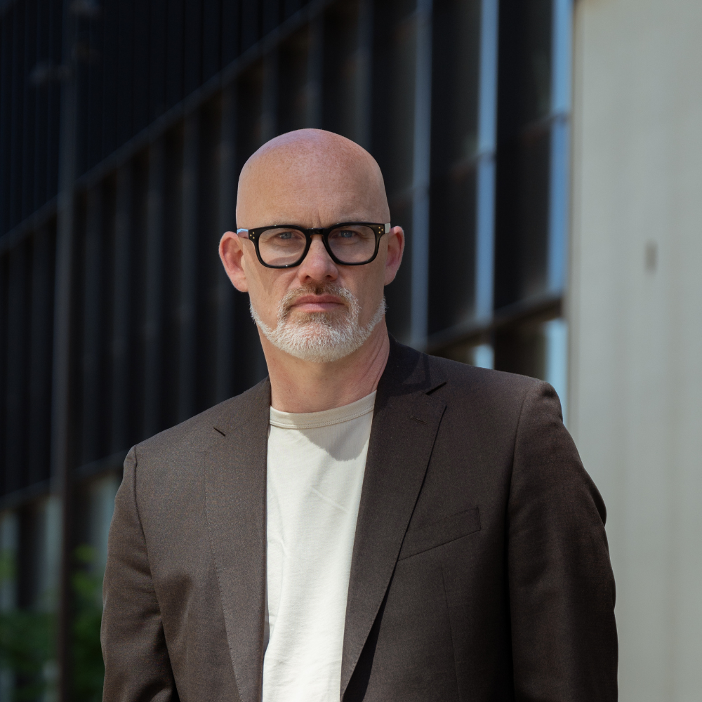 Bald man with glasses, wearing a brown blazer and white shirt, standing in front of a modern building.