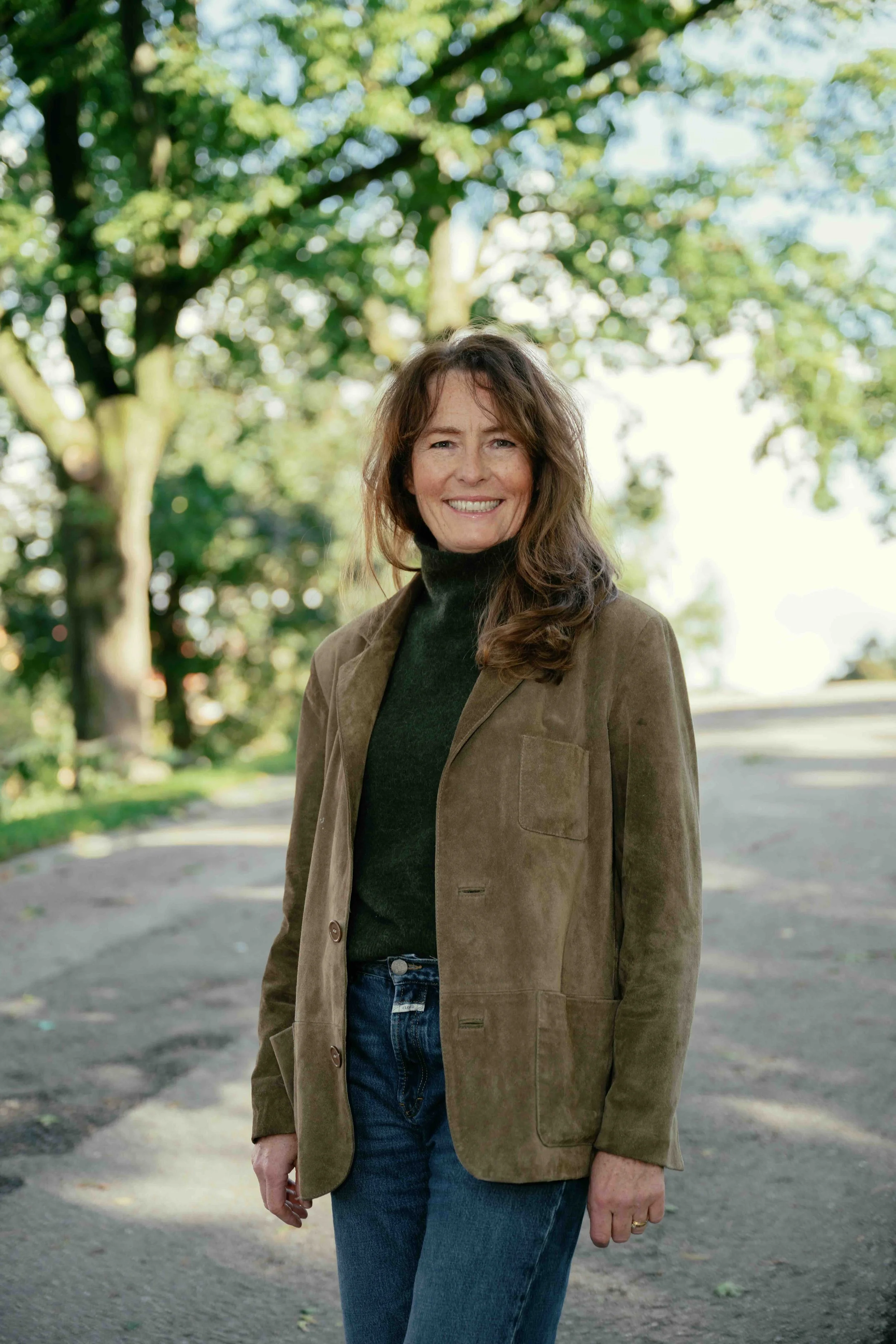 A woman with long brown hair smiling outdoors, wearing a black turtleneck, a brown blazer, and blue jeans, standing on a dirt path with green trees in the background.