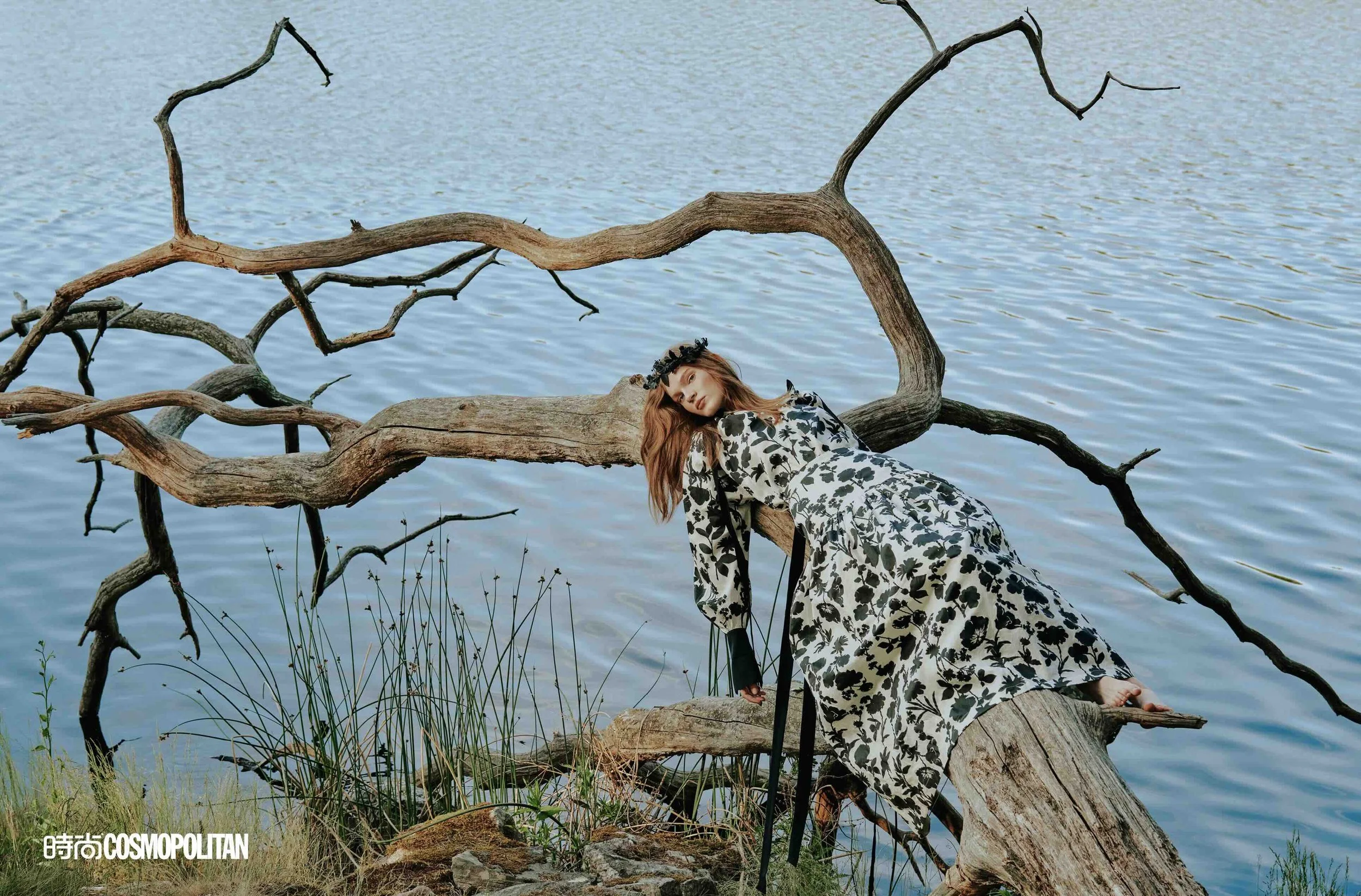 A woman wearing a long, floral dress with black and white patterns, lying on a fallen tree branch near a body of water, with the backdrop of a large, leafless tree limb arching over her.
