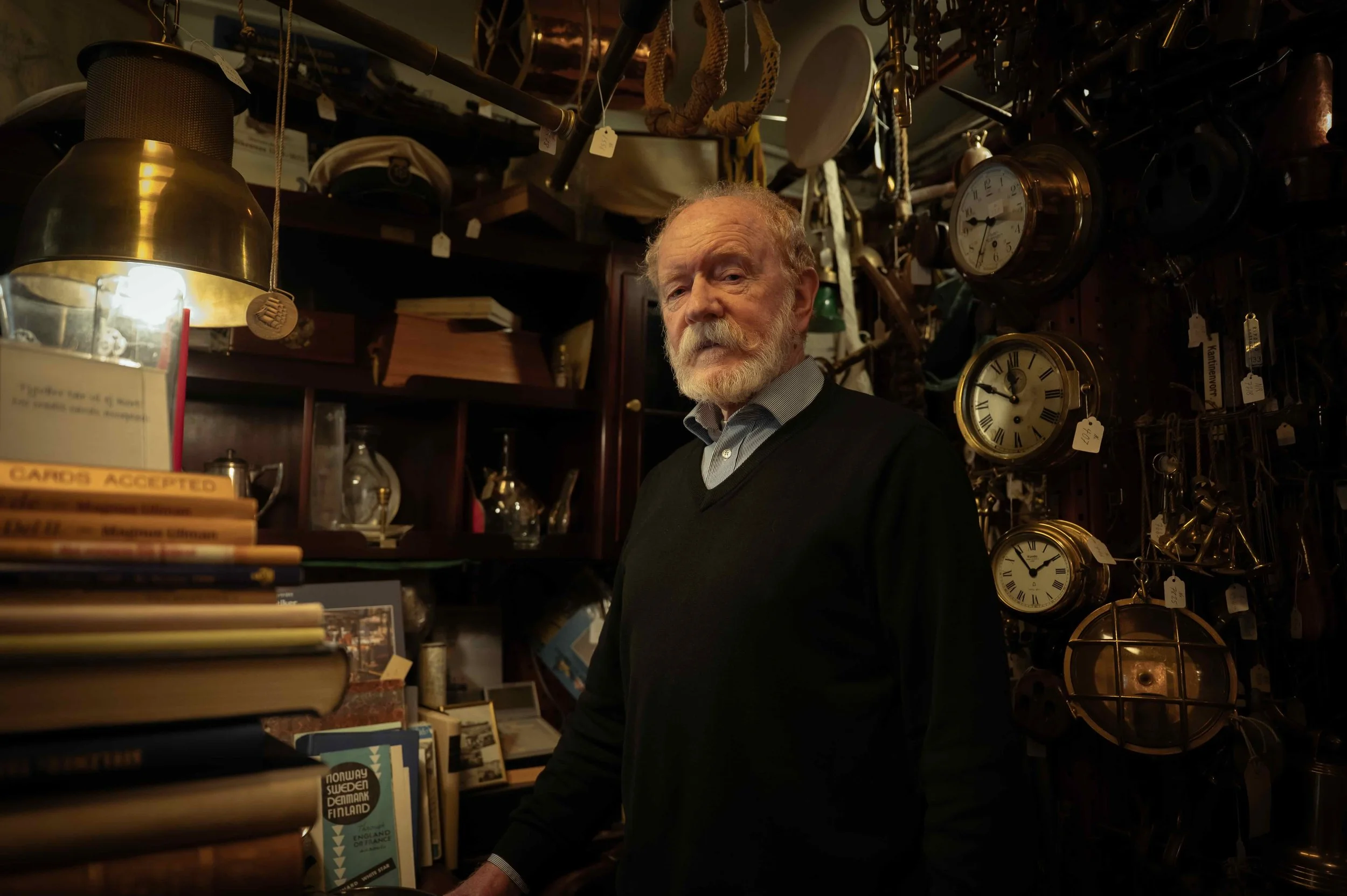 An elderly man with a white beard and mustache, wearing a black sweater and dress shirt, standing inside a cluttered antique shop surrounded by clocks, books, and various vintage items.