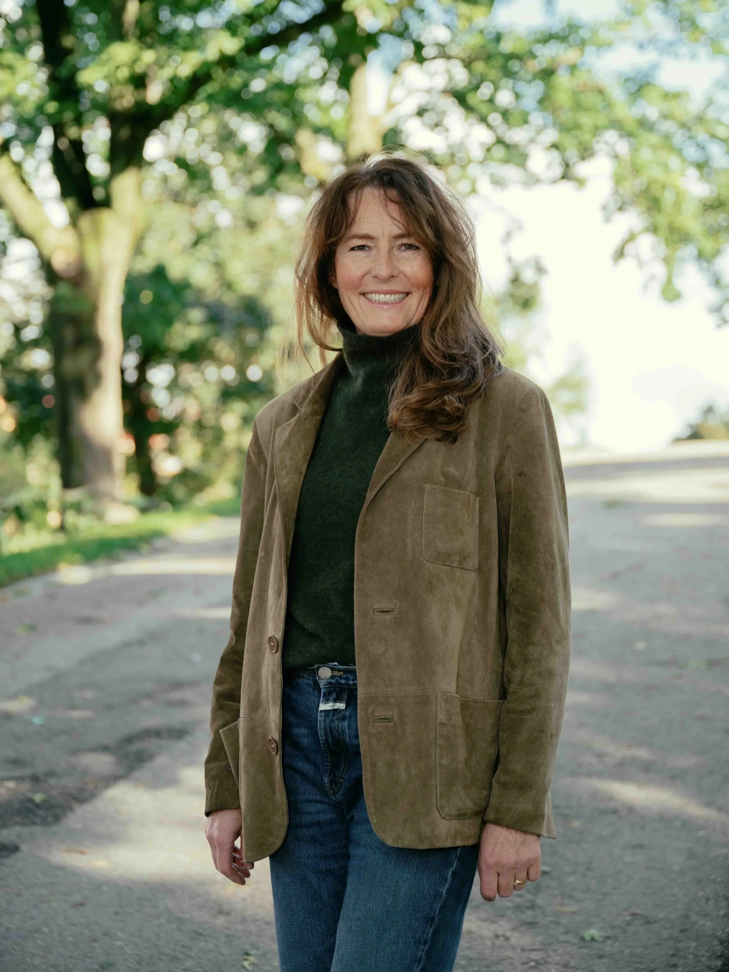 A woman with shoulder-length brown hair smiling outdoors in a park with green trees, wearing a brown blazer, black turtleneck, and blue jeans.