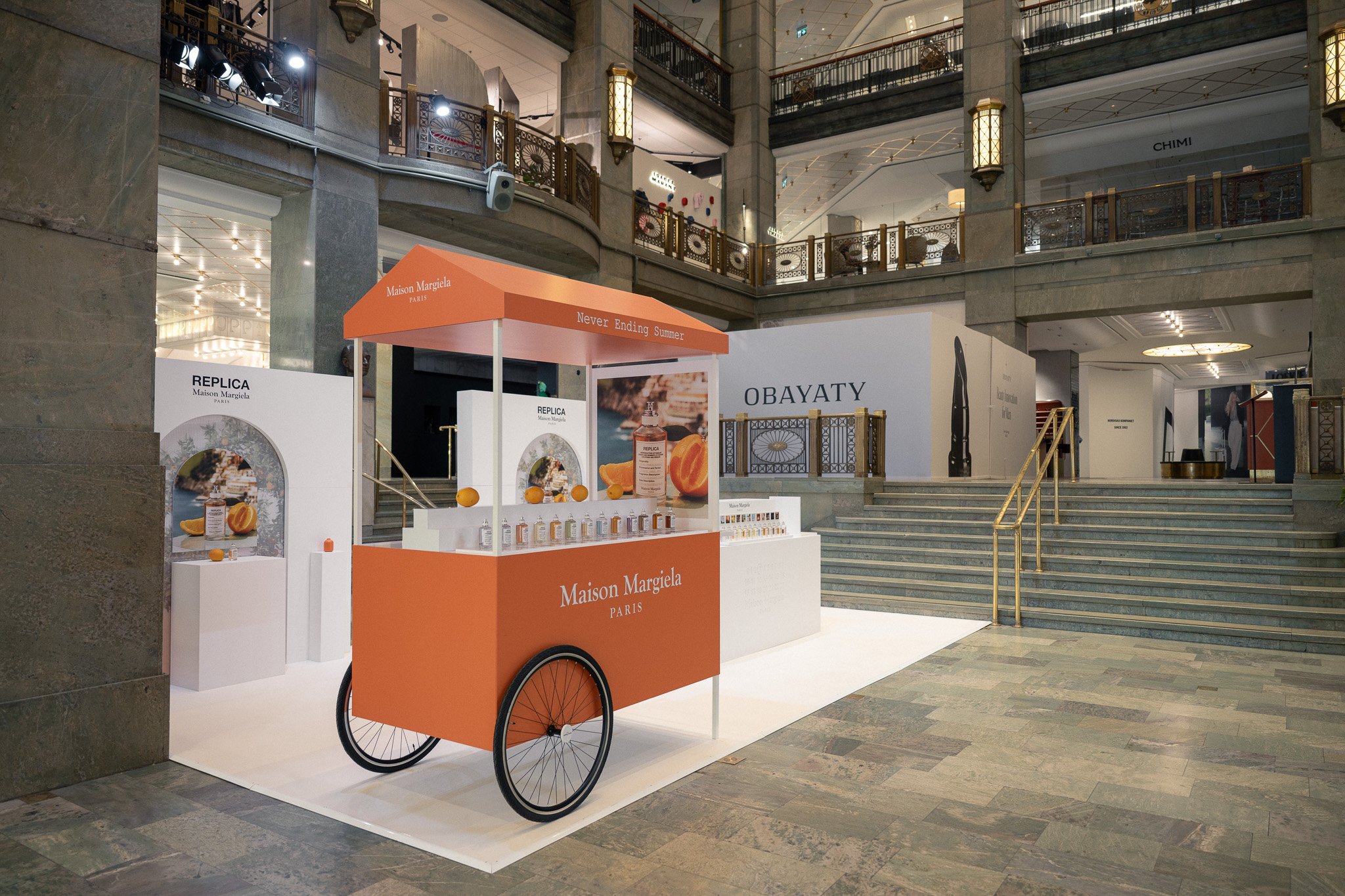An orange Maison Margiela Paris cart with a canopy labeled "Never Ending Summer" is set up in a shopping mall. The cart displays small bottles of perfume and oranges. The mall has marble stairs, ornate railings, and illuminated wall lanterns in the background.