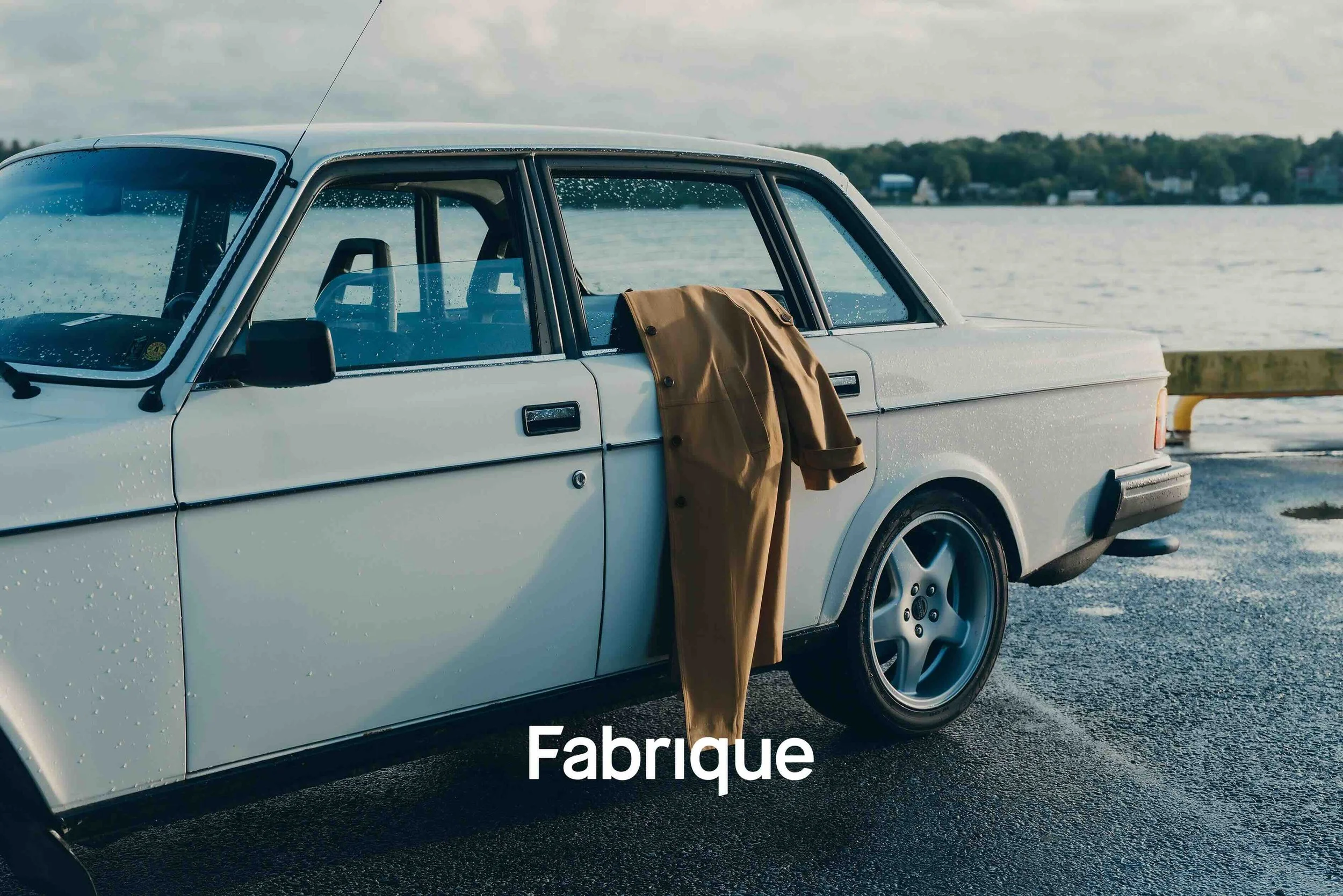Ett modereportage från Fabrique höst/vinter 2026 där en brun kappa visas upp vid en vit Volvo 240 i vintage-stil vid vattnet i Stockholm.