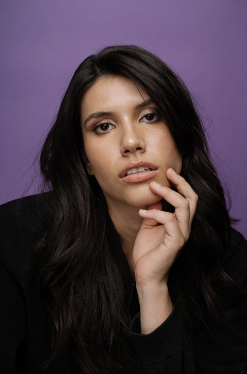 Portrait of a woman with long dark hair, posing with her hand near her face against a purple background.