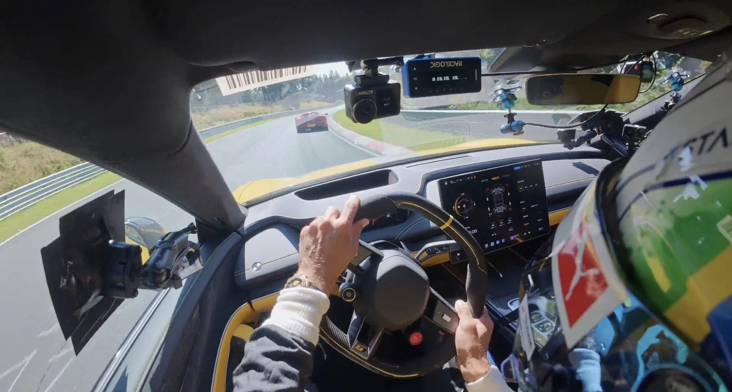View from the inside of a racing car, with a driver wearing a colorful helmet, racing on a track with another race car ahead and the driver's hand on the steering wheel.
