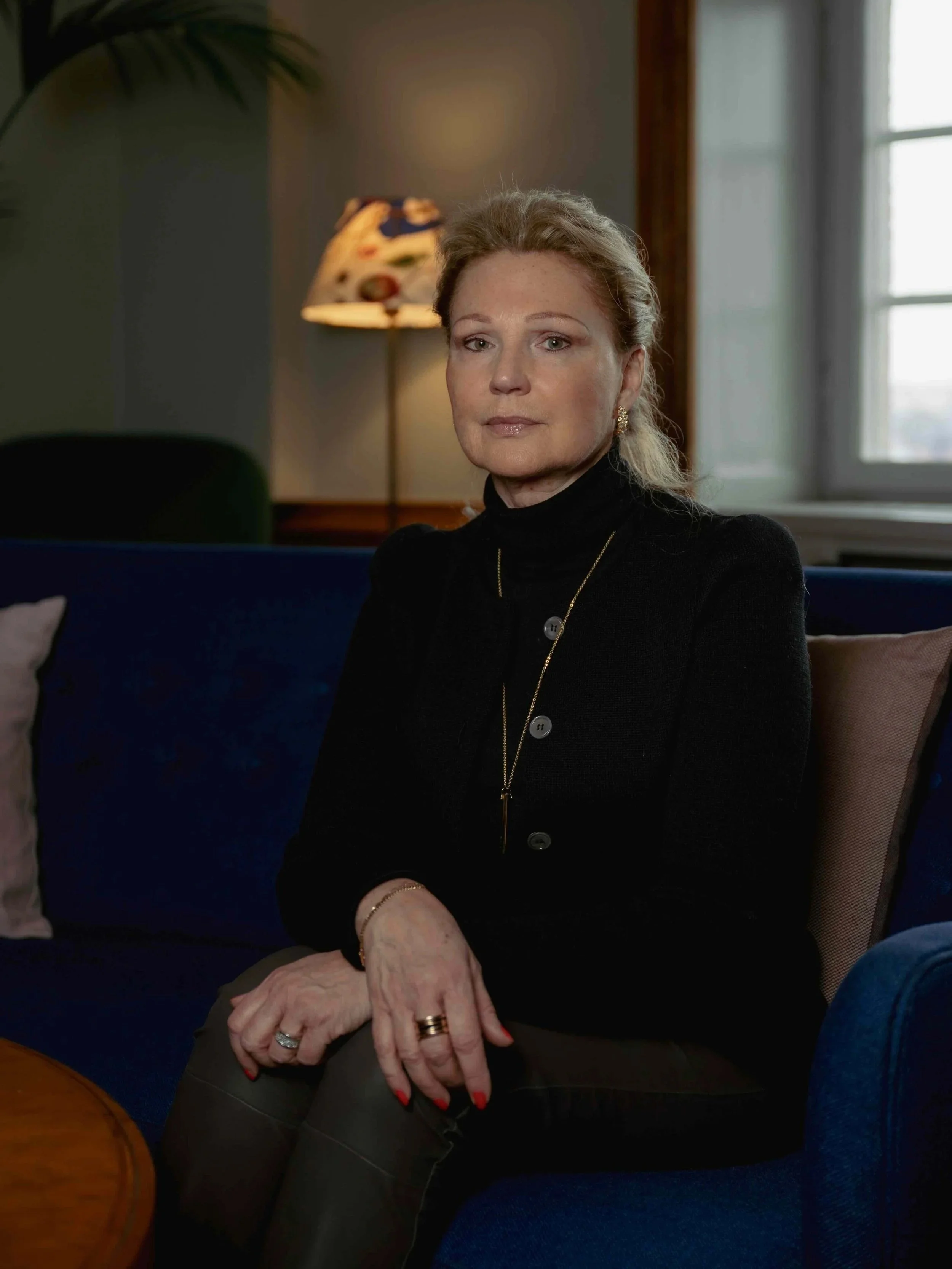 A woman with blonde hair sitting on a blue couch in a living room. She is wearing a black turtleneck, gold jewelry, and has groomed nails. The background includes a window with white curtains and a lamp with a colorful abstract shade.