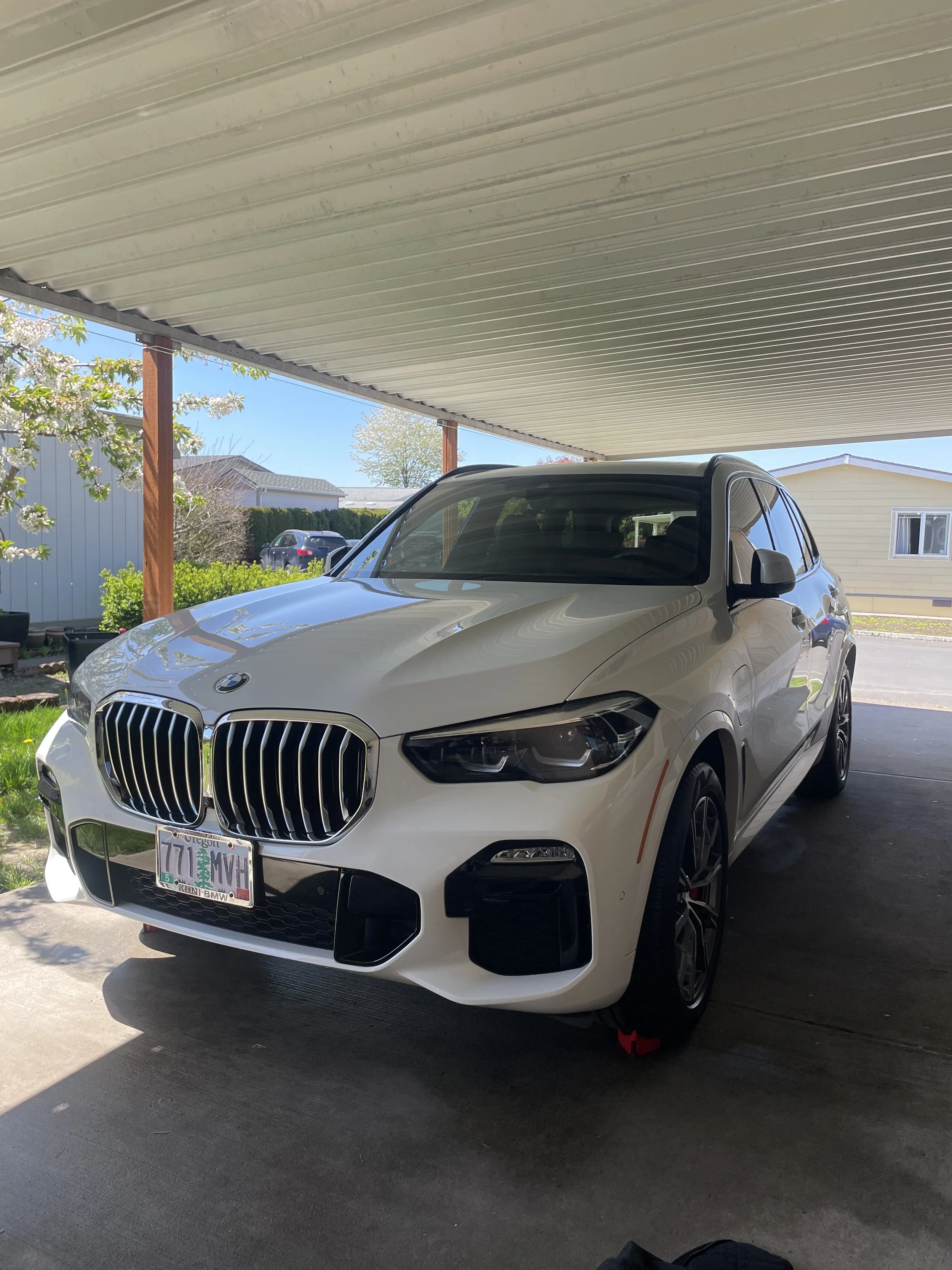 White BMW SUV parked in a covered driveway with a garden and house in the background.