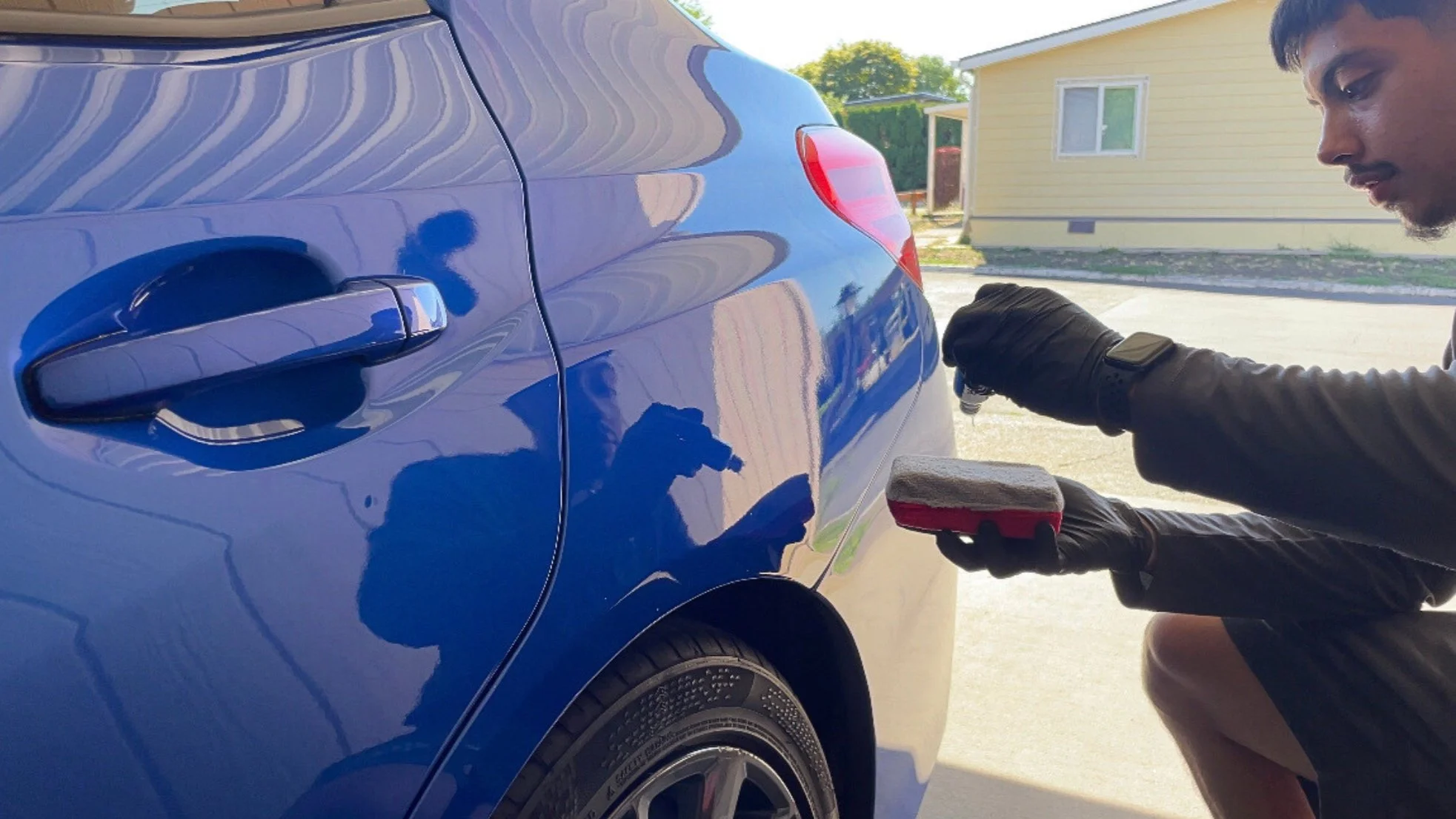 A man in black gloves is polishing a blue car with a red and gray sponge on a sunny day, with a yellow house and trees in the background.