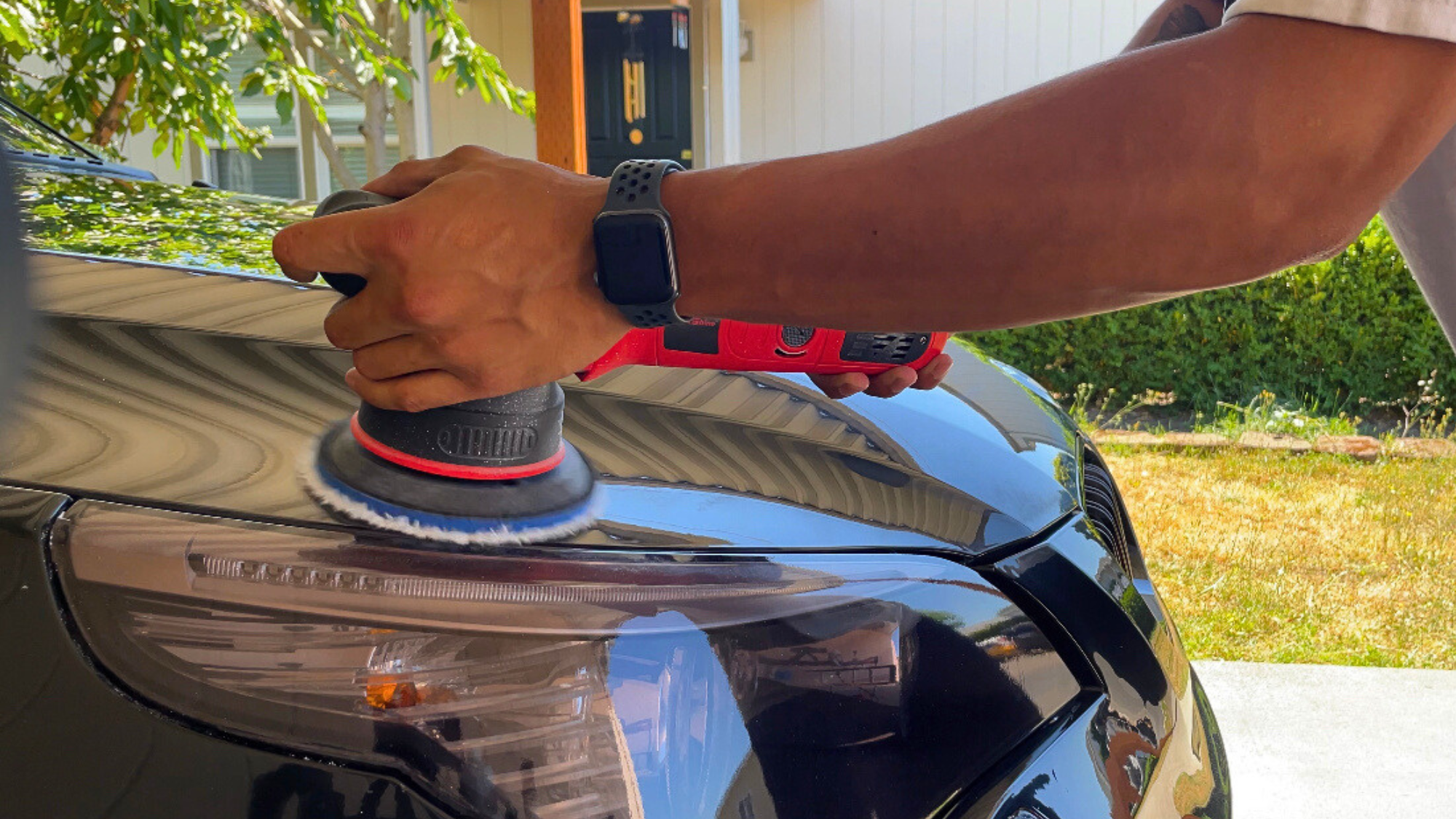 Person polishing the black car's headlight with a red and black polishing machine outside, wearing a black wristwatch, with a porch and trees in the background.