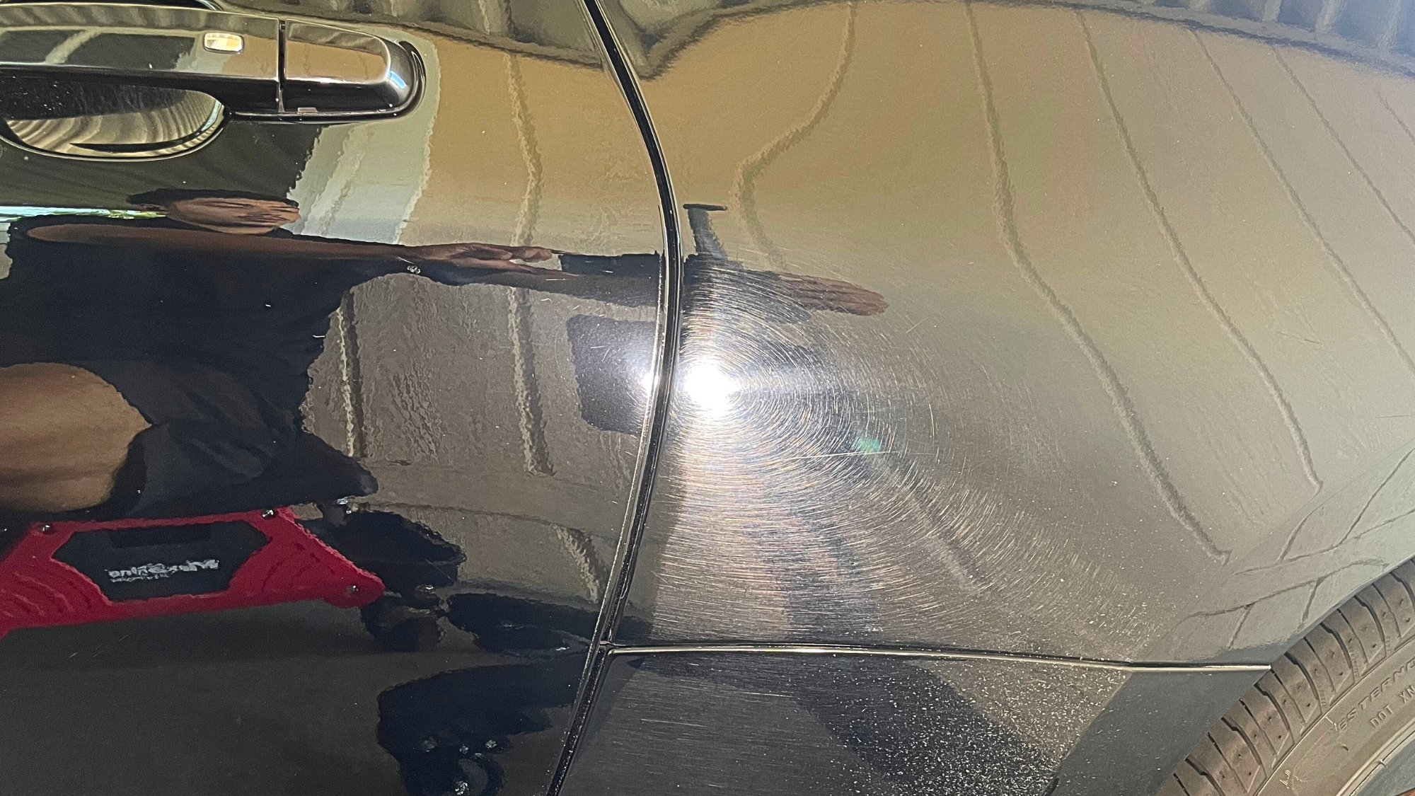 Close-up of a black car's dented and scratched passenger side door with visible swirl marks, next to the wheel and tire.