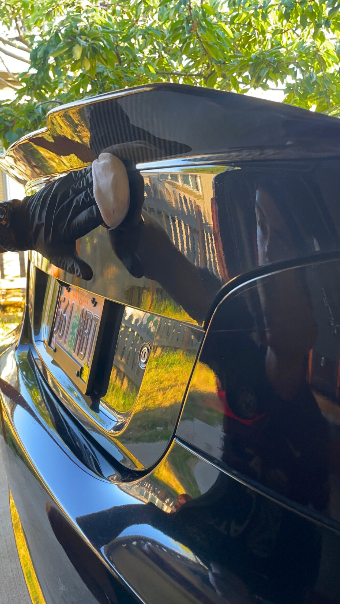 A person wearing gloves cleaning the back of a black car with a polishing pad, with a tree and a fence in the background.