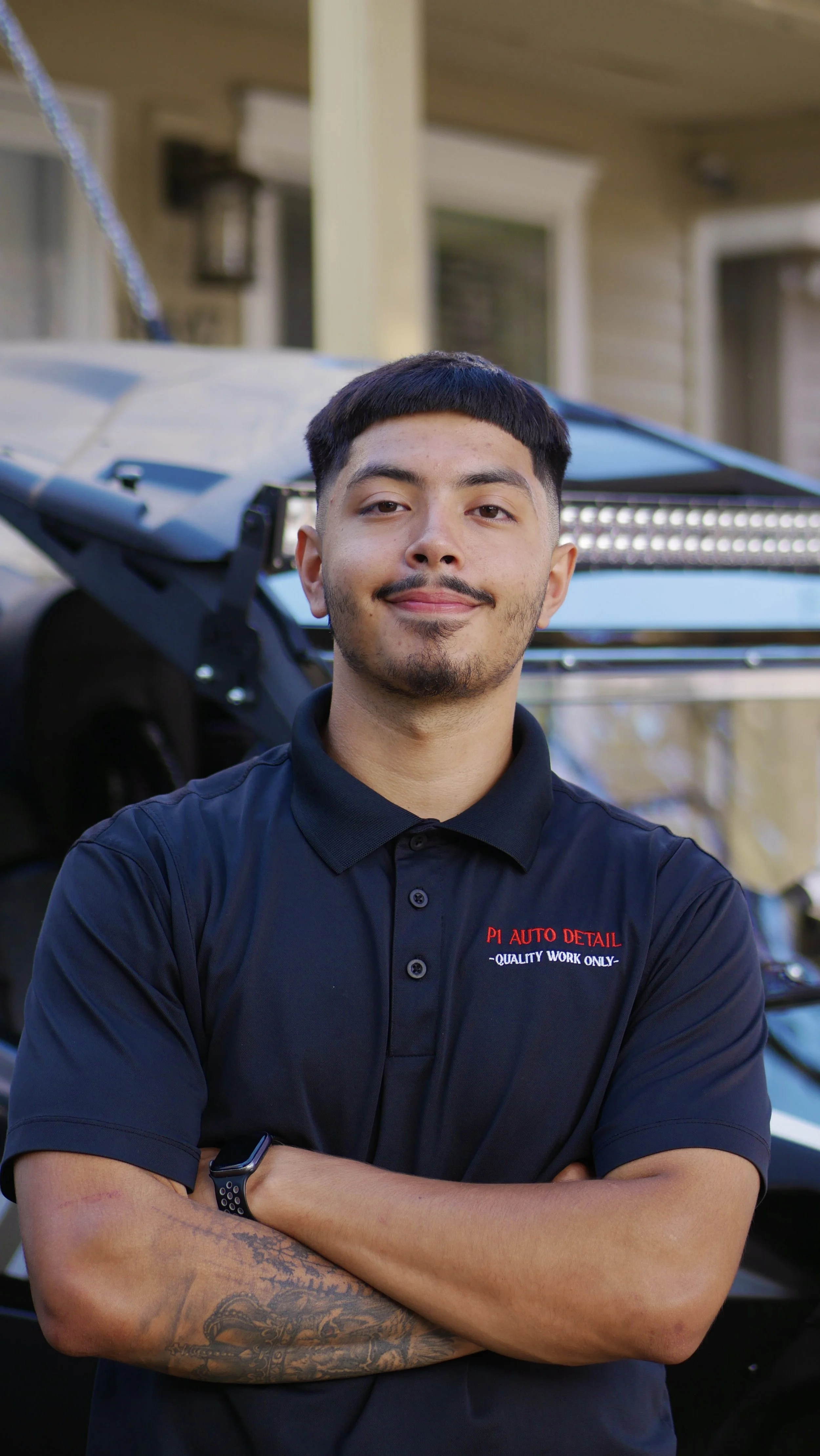 A man with tattoos on his left forearm, wearing a black polo shirt with red and white text, standing with arms crossed in front of a black sports car and a light-colored house with a porch.