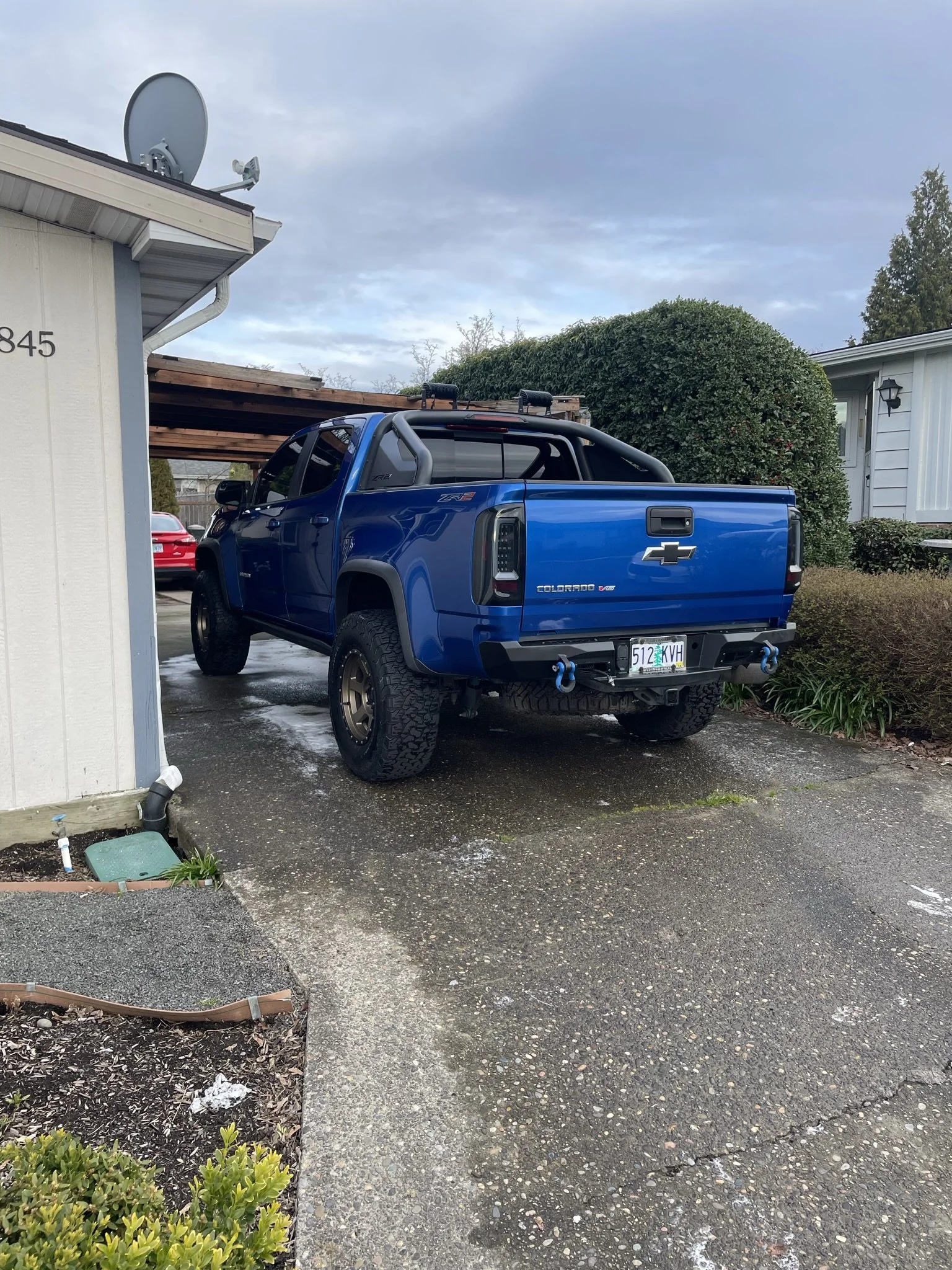 A blue Chevrolet Colorado pickup truck parked on a wet driveway next to a white house with the number 845 on the wall. The truck has off-road tires, black accents, and a custom roll cage with blue tow hooks.