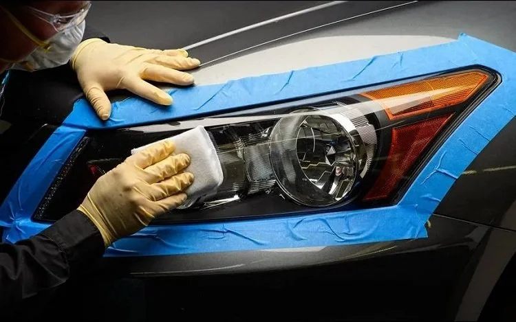 A person wearing yellow gloves is cleaning or polishing a car headlight with a cloth, with blue painter's tape around the headlight for protection.