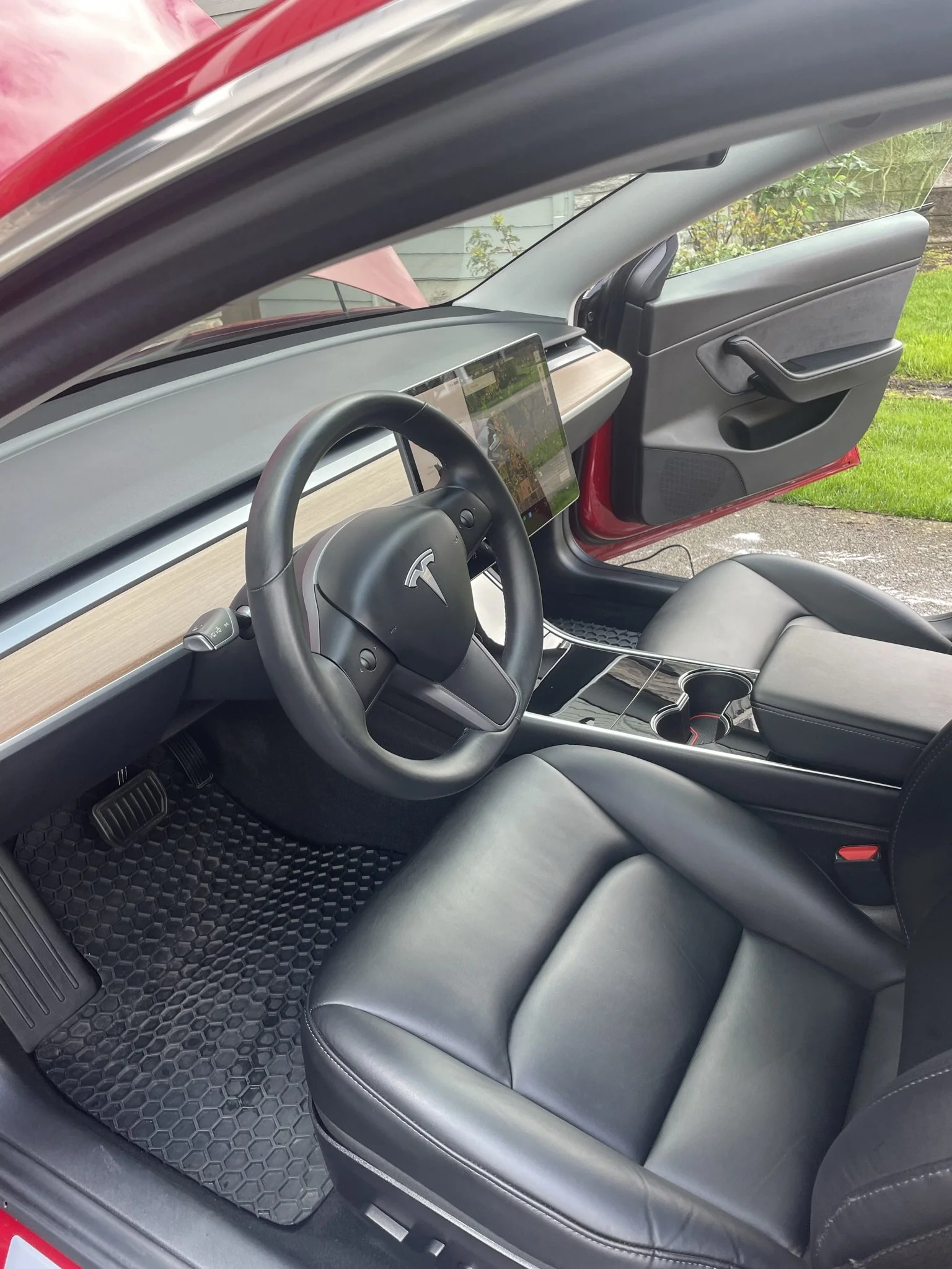 The interior of a Tesla electric vehicle, showing the steering wheel, dashboard, touchscreen display, and black leather seats, with a view of the outside grass and sidewalk through the open door.