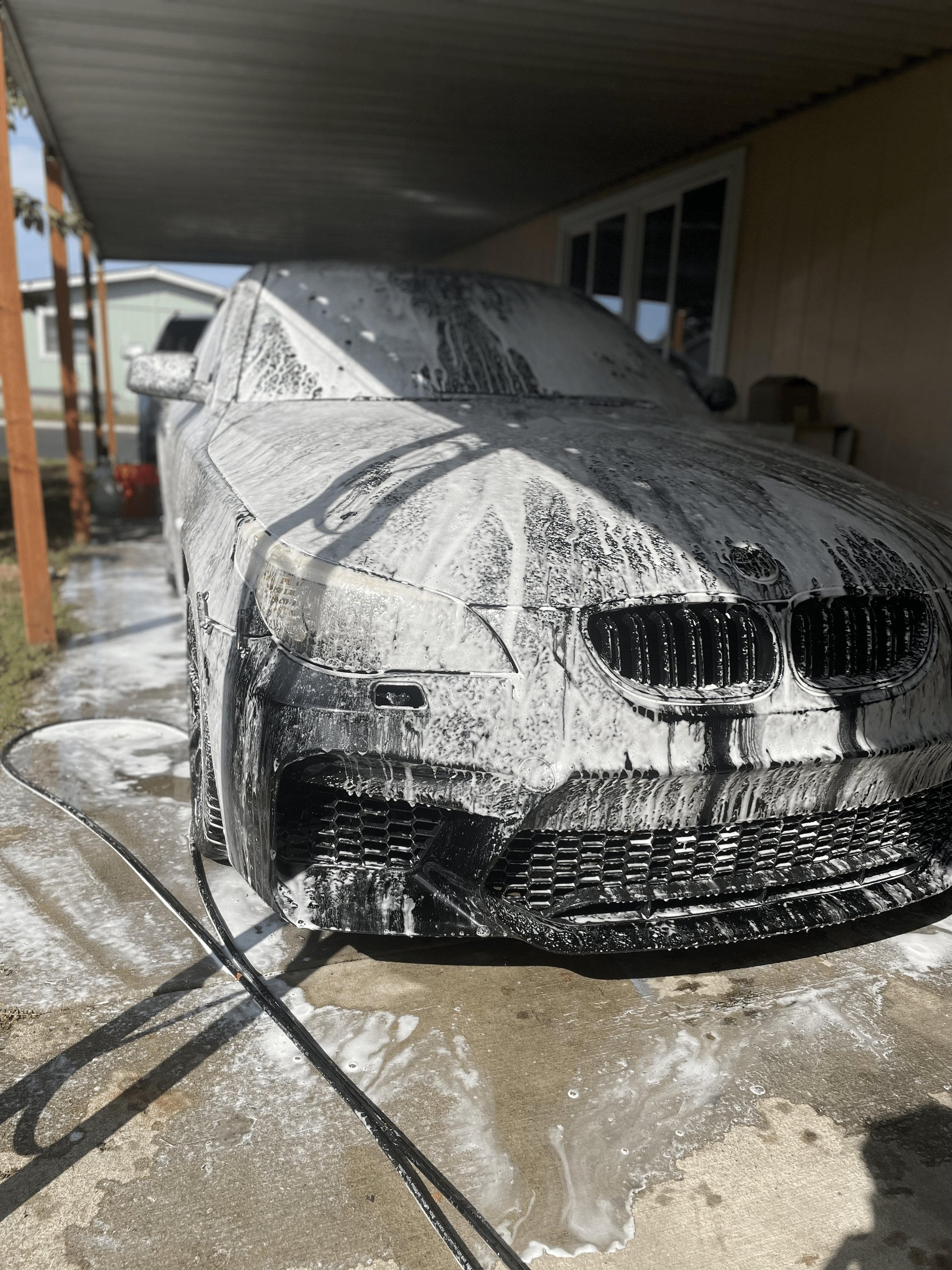 White car being washed with soap and foam, parked under a carport on a concrete driveway.