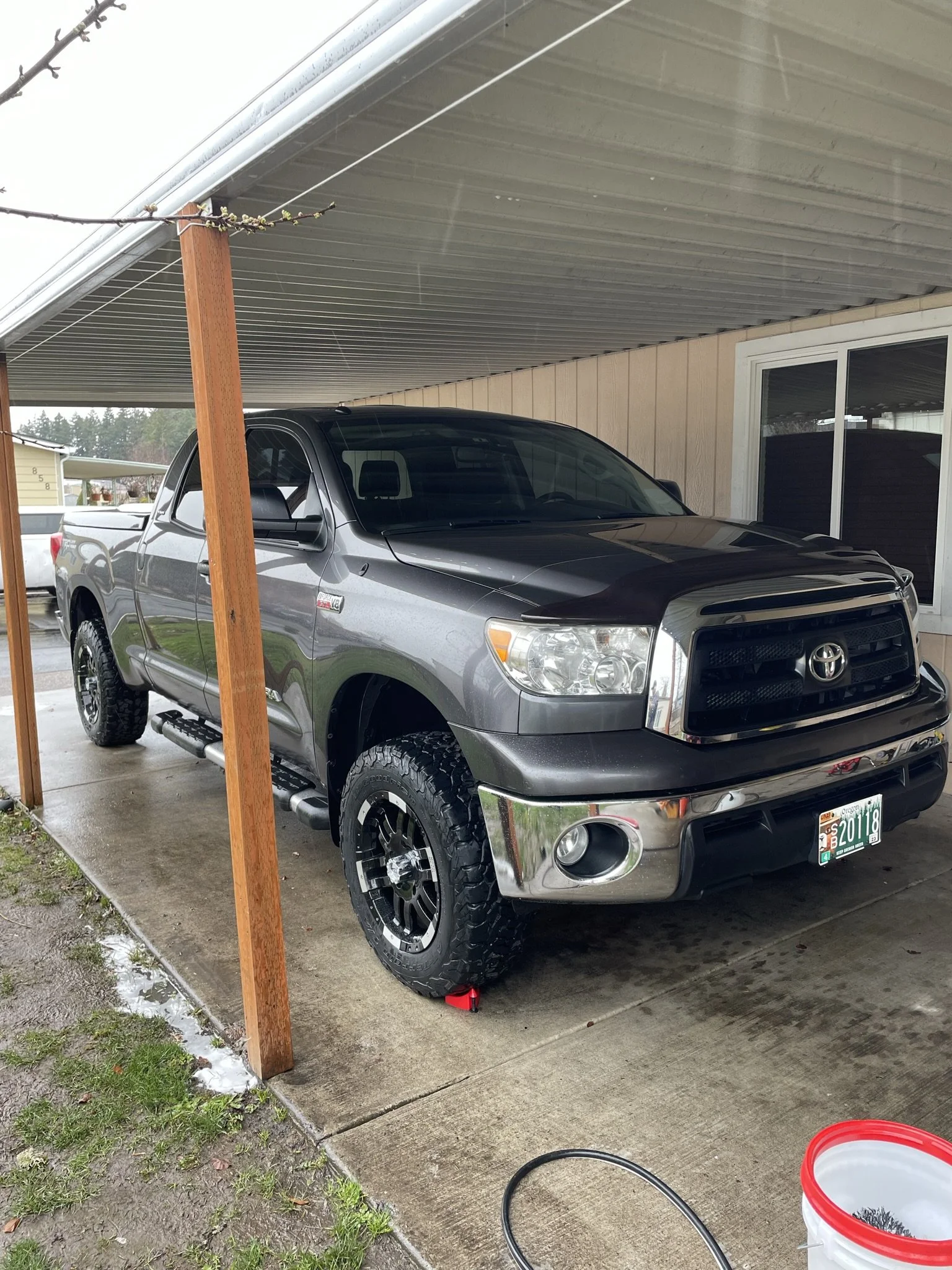 A gray Toyota pickup truck with off-road tires parked under a carport attached to a house.