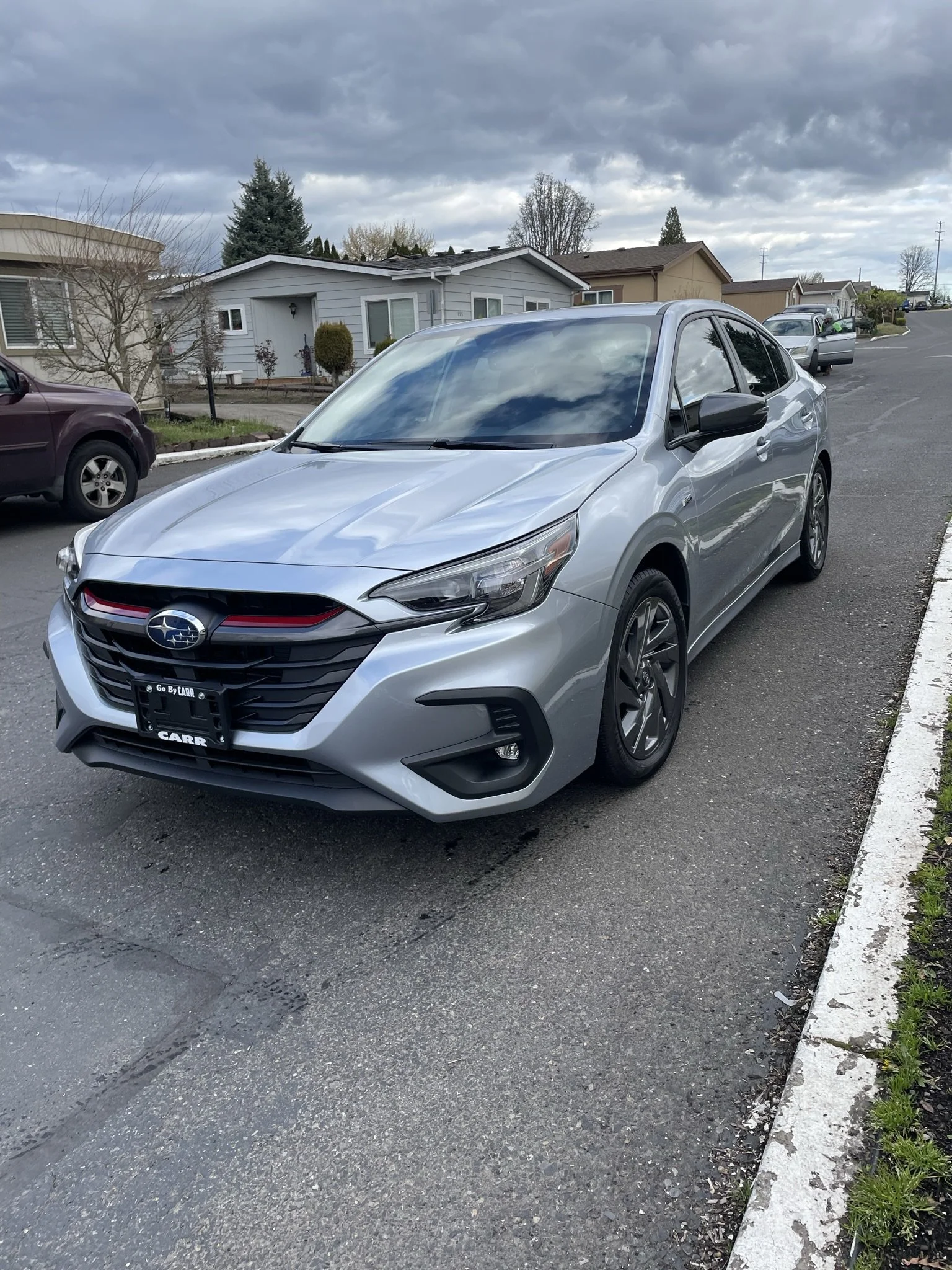 A gray sedan parked on the side of a residential street with houses and other cars in the background, under a cloudy sky.
