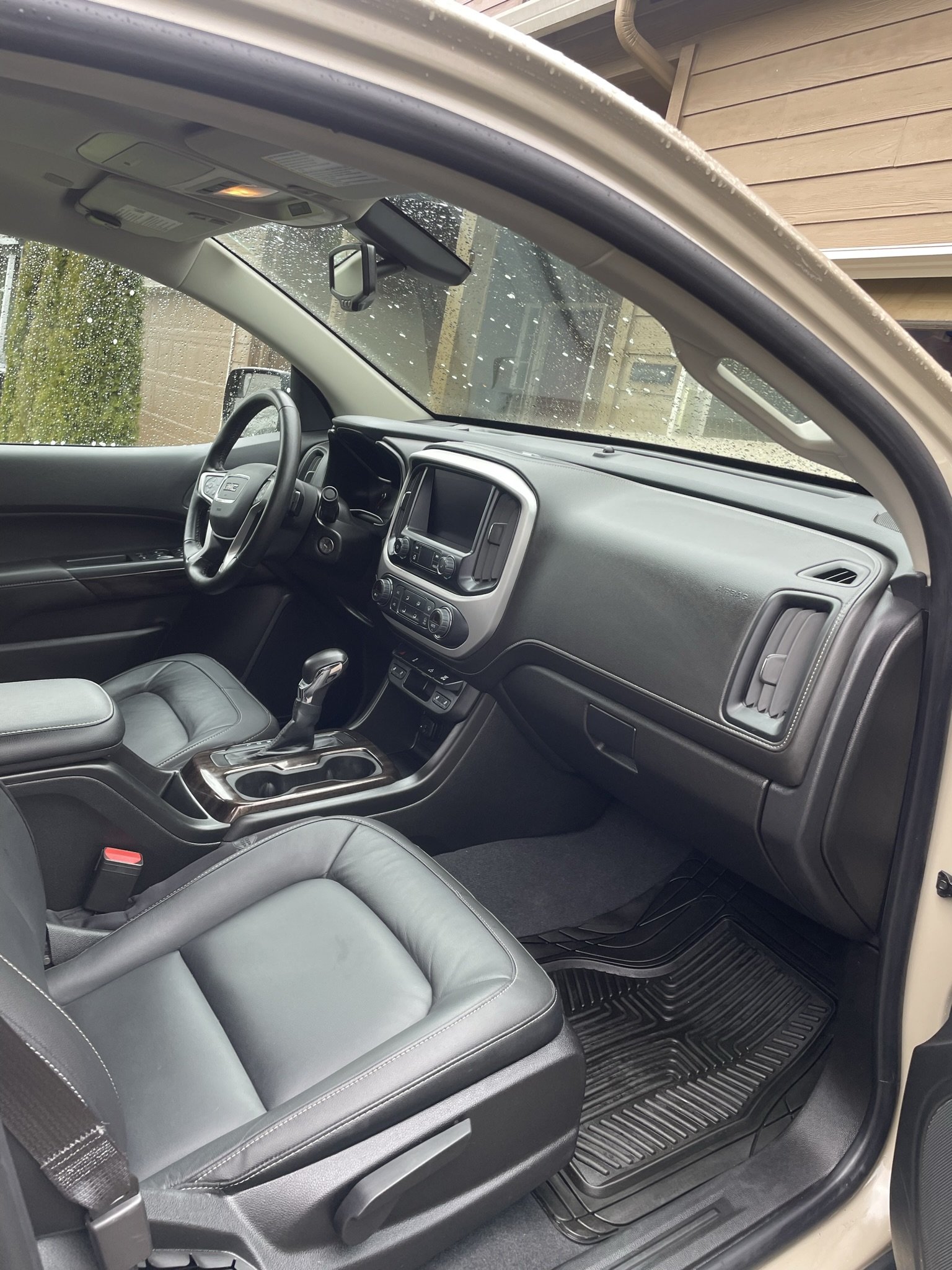 Interior of a vehicle showing the dashboard, steering wheel, front seats, and floor mats, with rain on the windshield.