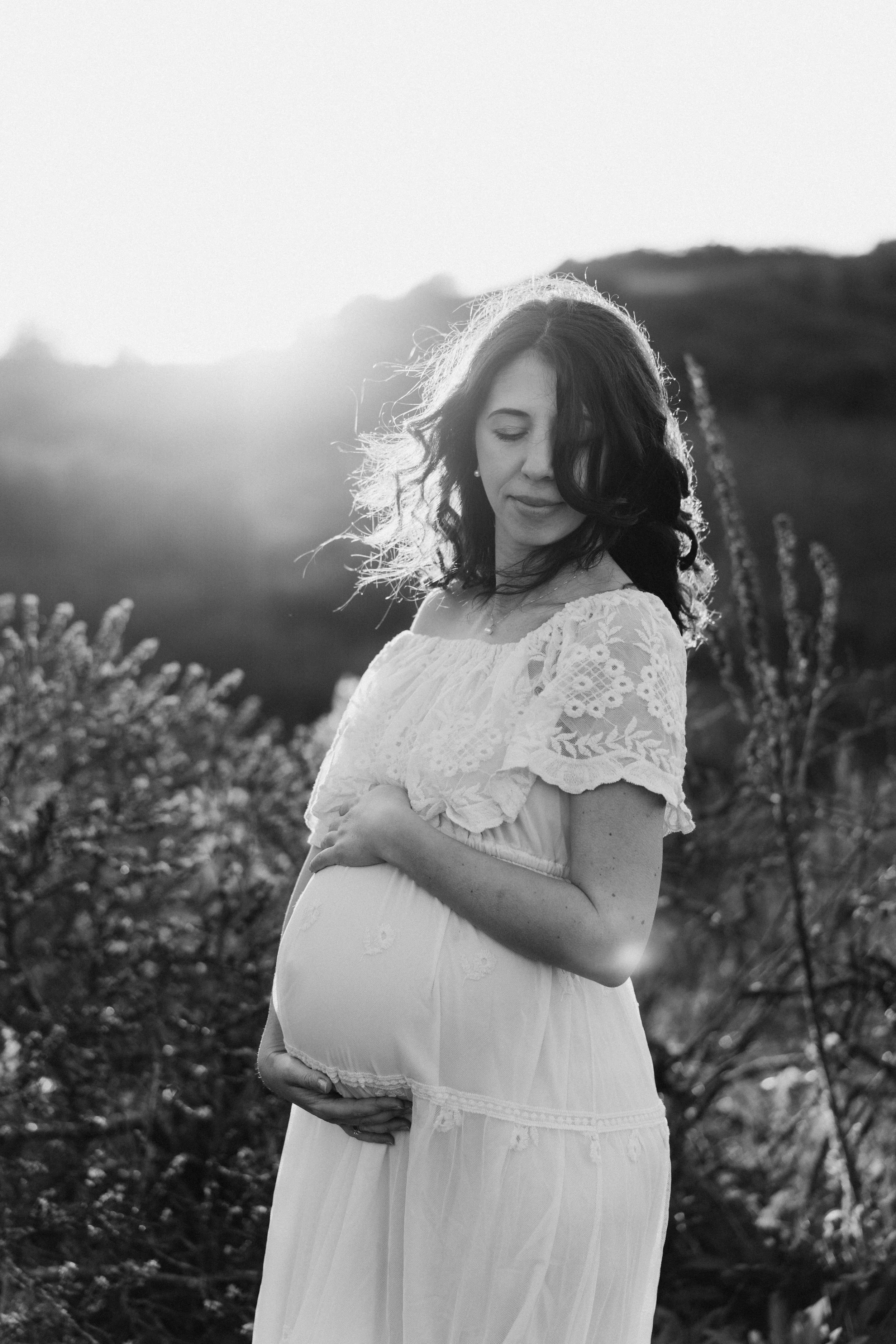 pregnant woman in a white dress gently holding her baby bump during an outdoor maternity photshoot