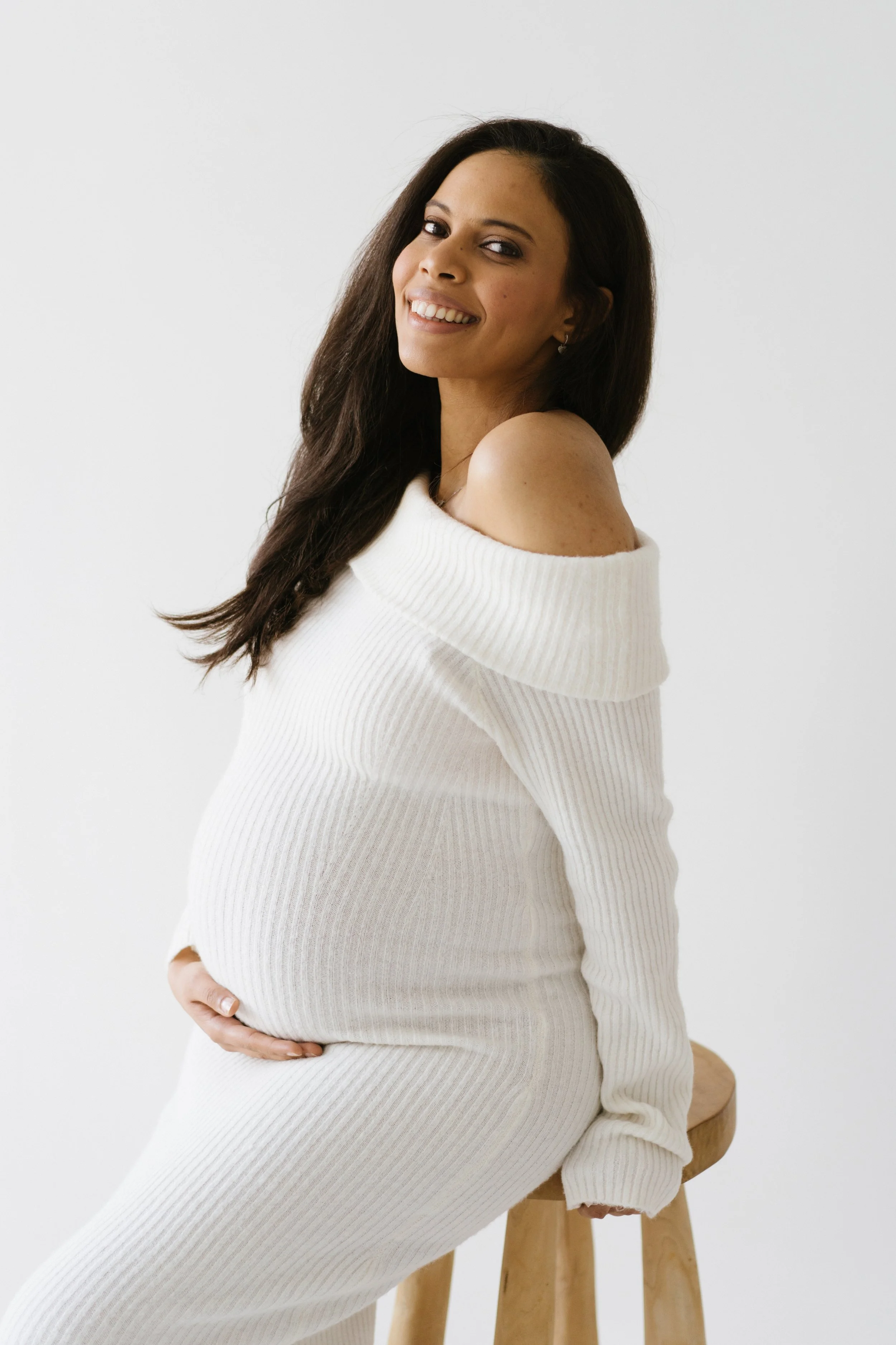 smiling pregnant woman in a white off-shoulder dress, seated and gently holding her baby bump during a maternity photoshoot