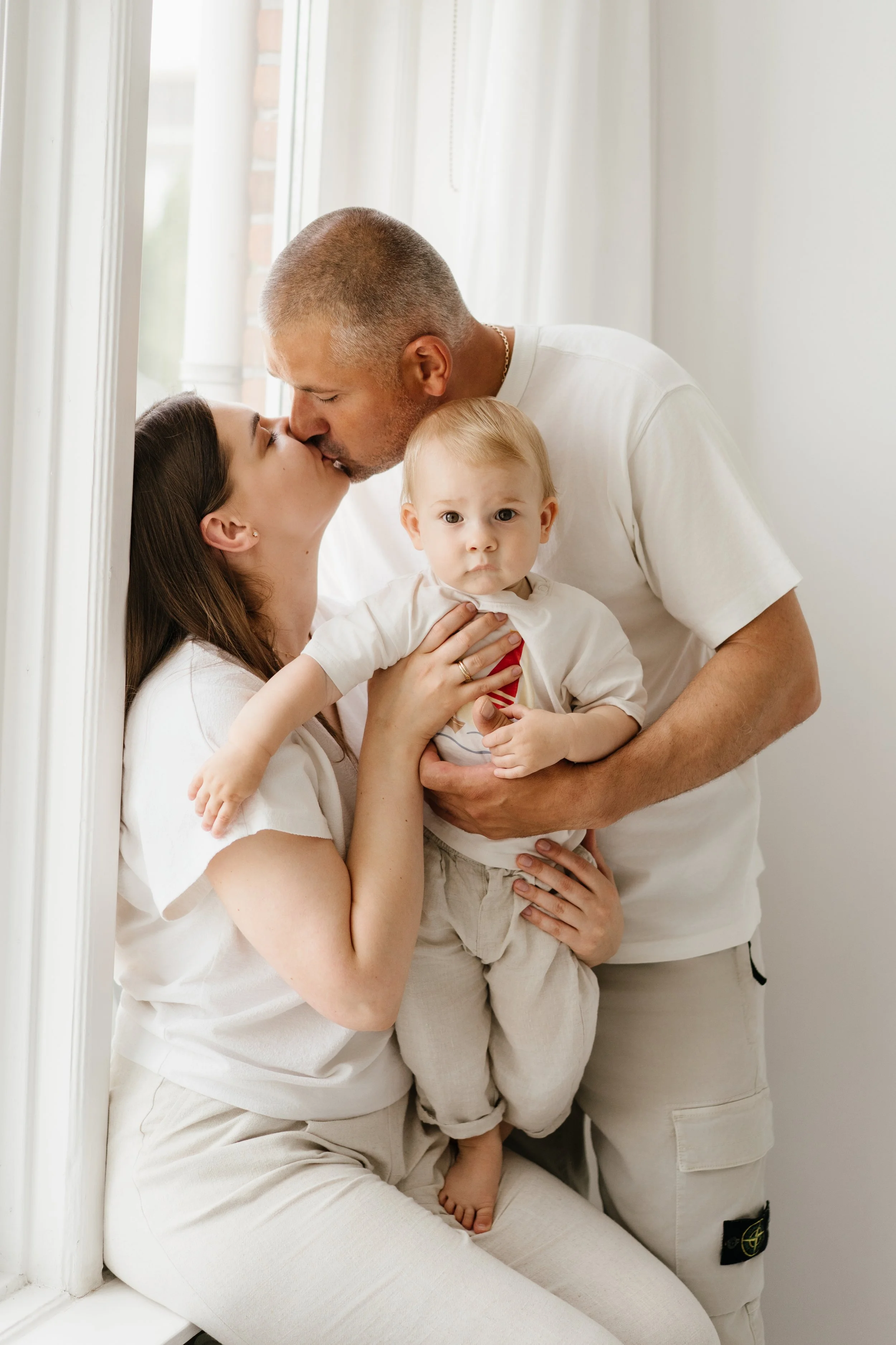 parents sharing a kiss while holding their young child during a warm, natural family photoshoot