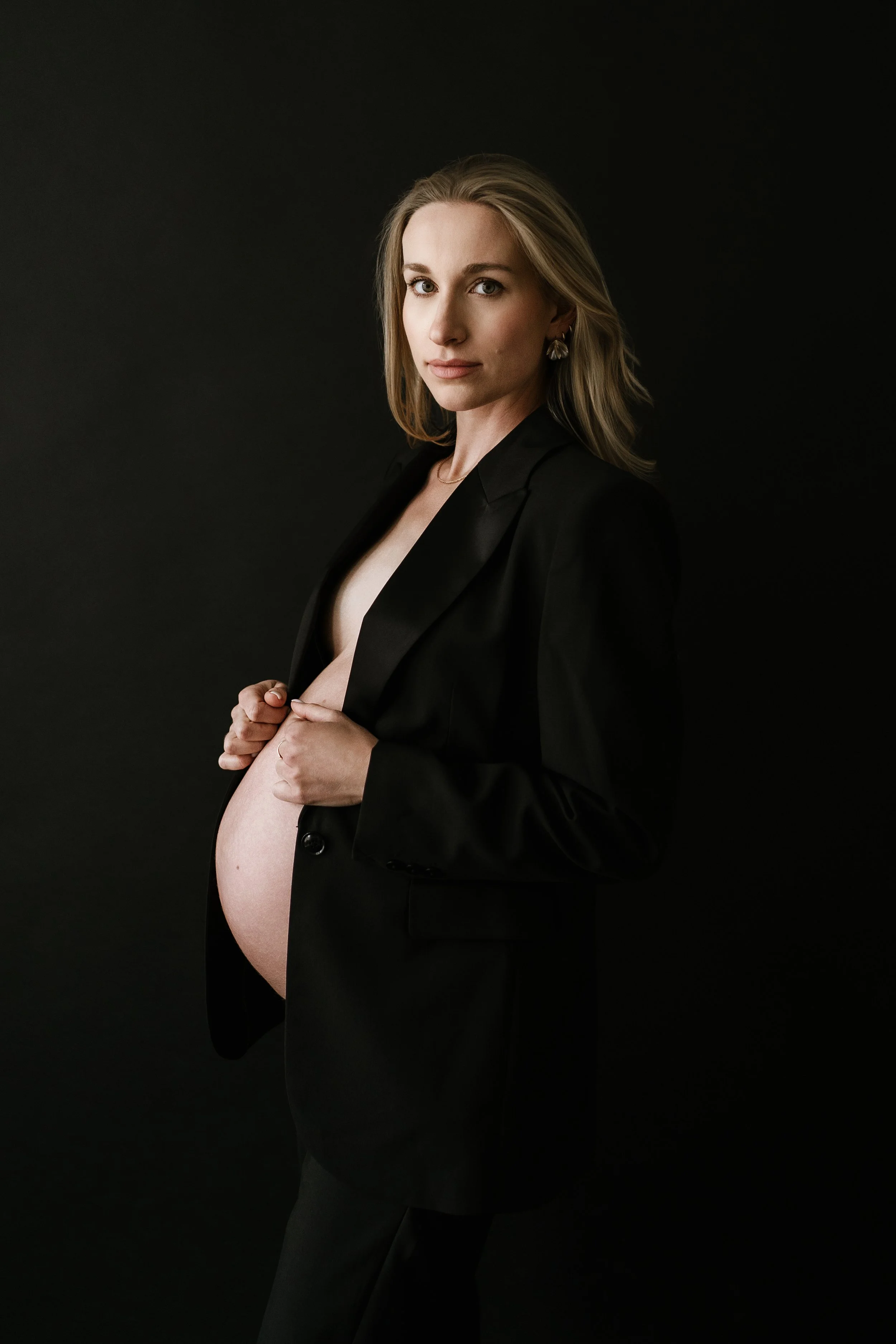 pregnant woman in a lack blazer posing against a dark background, gently holding her exposed baby bump during a maternity photoshoot