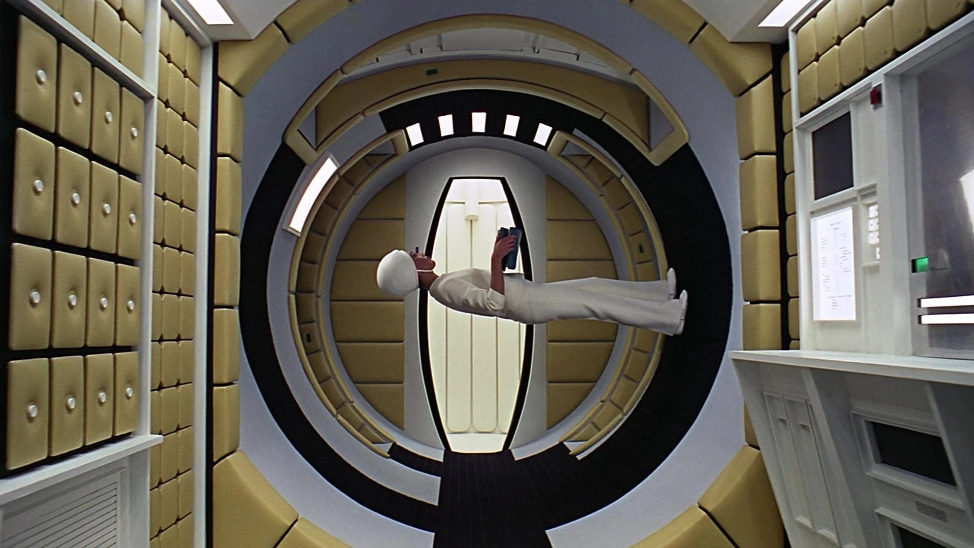A person in a white outfit and wig within a circular space station corridor, holding a tablet device.