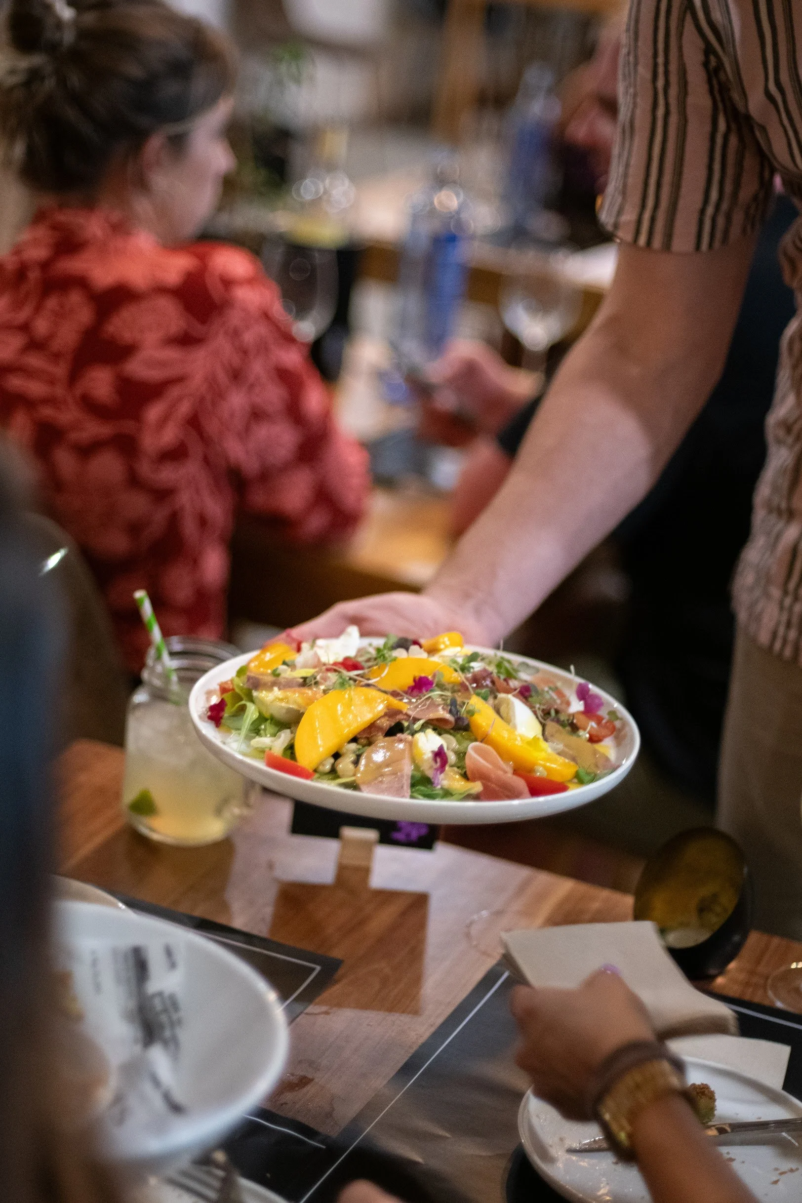 Person handing a colorful salad to another person at a restaurant table.