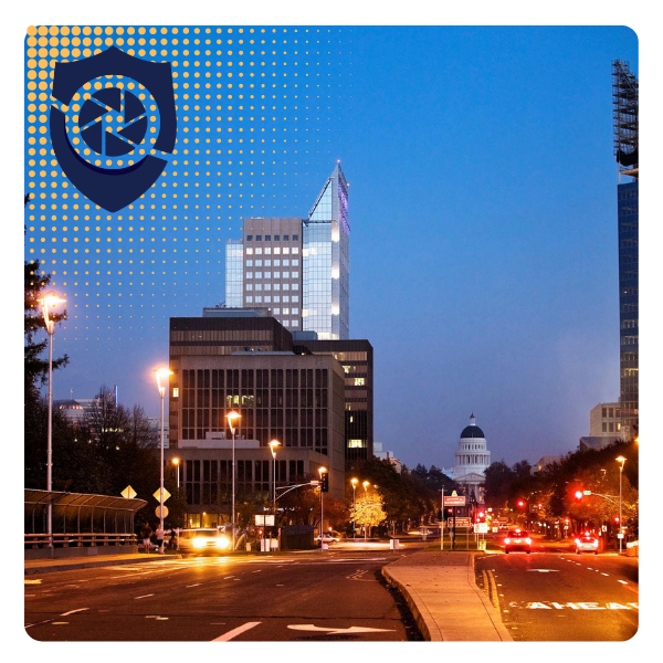 City street scene in evening with tall modern buildings, traffic, and the California State Capitol building visible in the background in Sacramento.