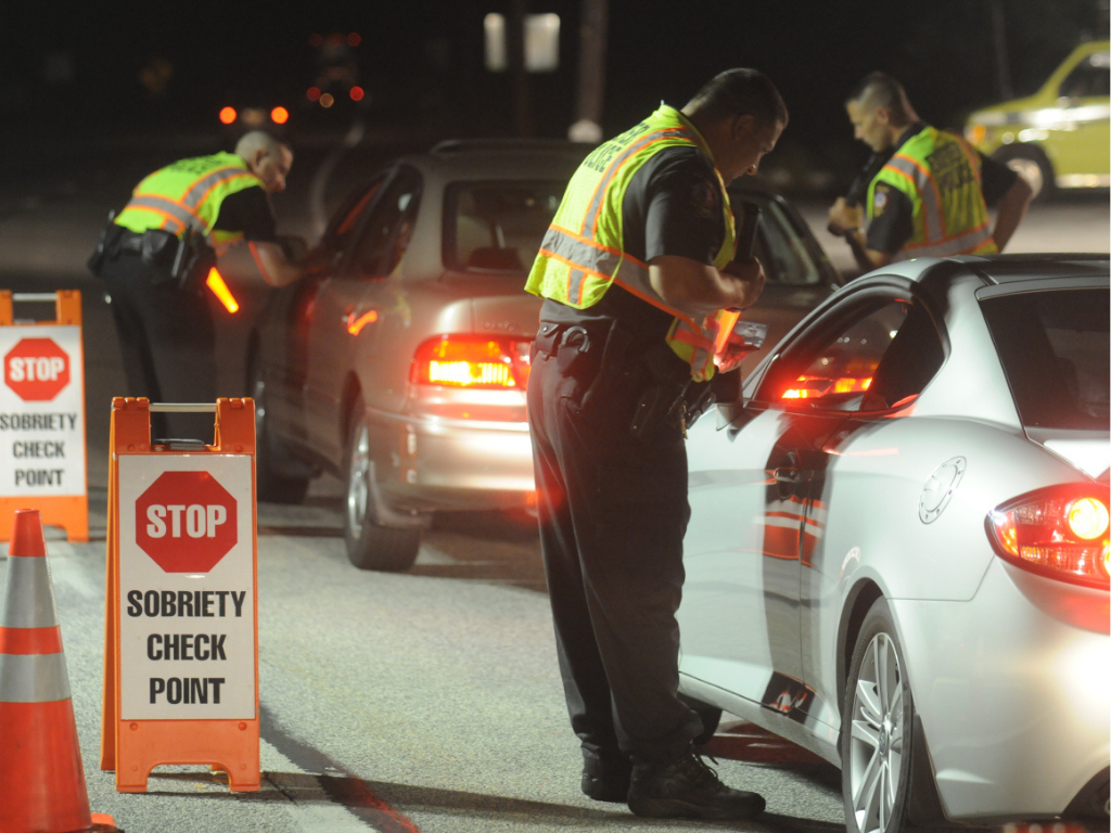 Police officers conducting a sobriety check on drivers at night on a road, with signs that read "STOP SOBRIETY CHECK POINT" and traffic cones.