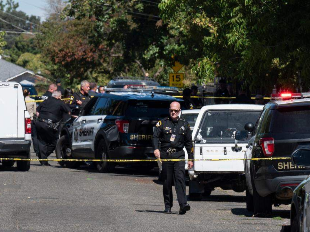 Police officers and sheriff vehicles at a crime scene, with yellow caution tape and trees in the background.