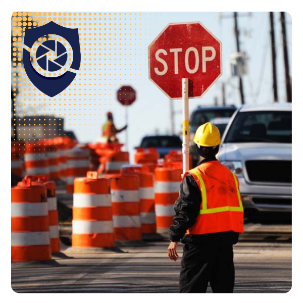 Construction worker in safety vest and helmet standing near a stop sign, with orange and white traffic barrels on a city street.