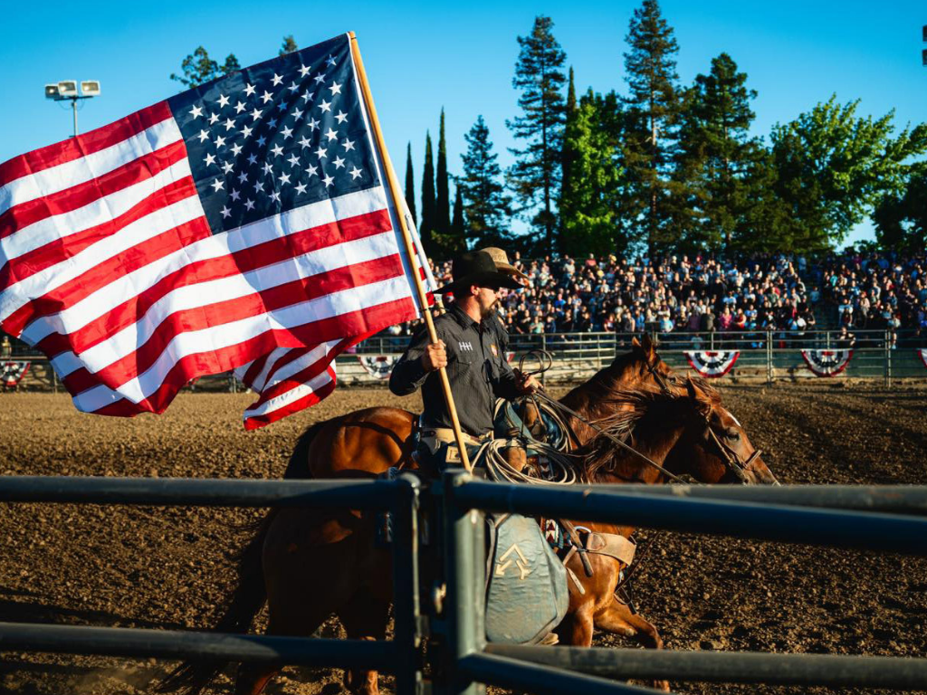 A man on horseback holding an American flag at a rodeo with a crowd in the background.