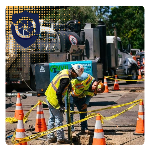 Two construction workers in safety vests and helmets working near orange traffic cones and yellow caution tape, with a large industrial truck and a utility vehicle in the background.