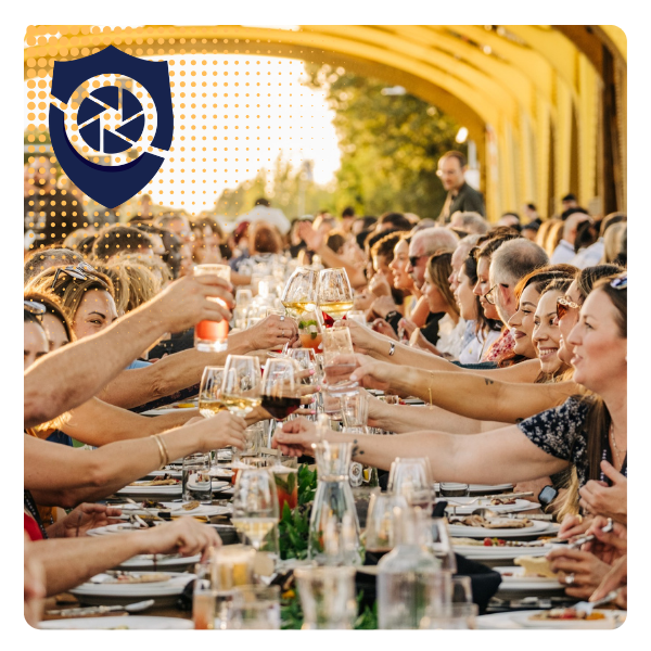 People gathered at long outdoor banquet table raising glasses for toast during a celebration. A yellow bridge or structure is in the background, with sunlight filtering through trees.