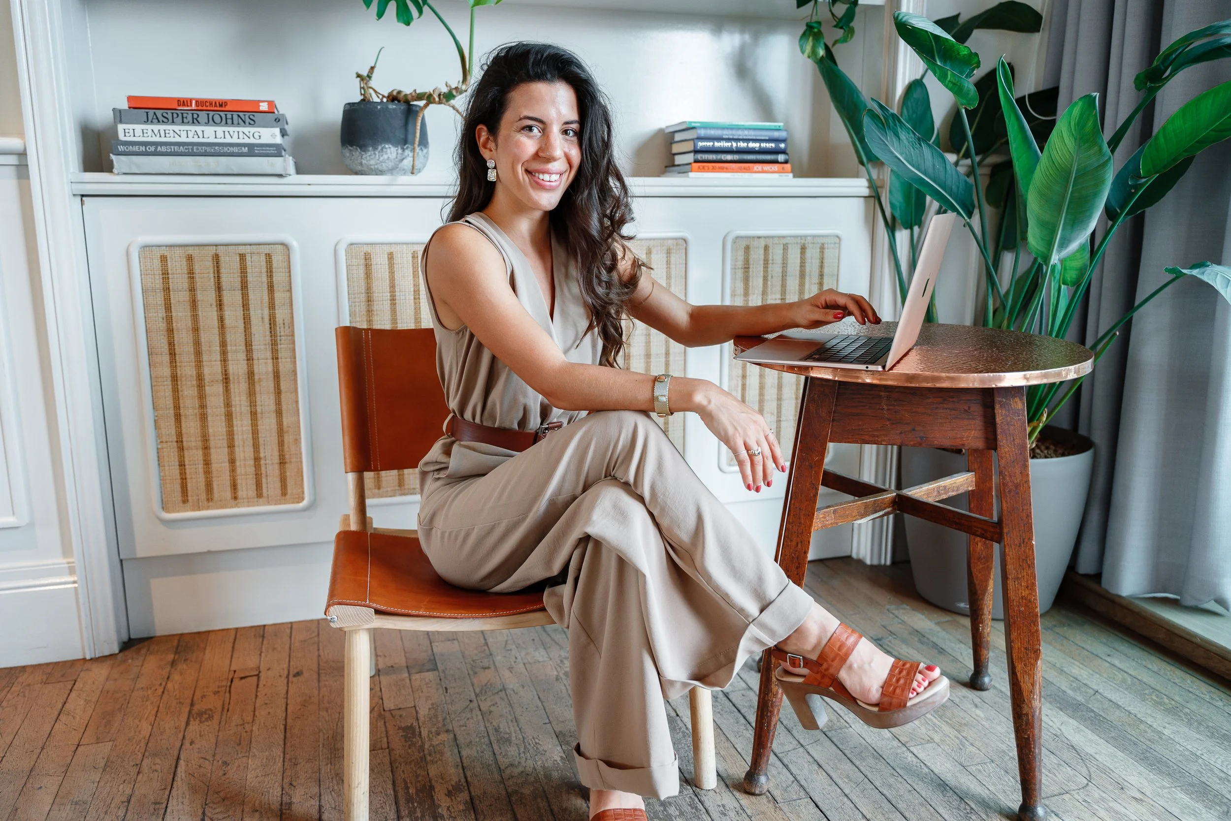 A woman with long dark hair, wearing a beige sleeveless jumpsuit and brown high-heeled sandals, is sitting on a wooden chair in a bright room. She has a laptop on a wooden side table next to her and is smiling at the camera.