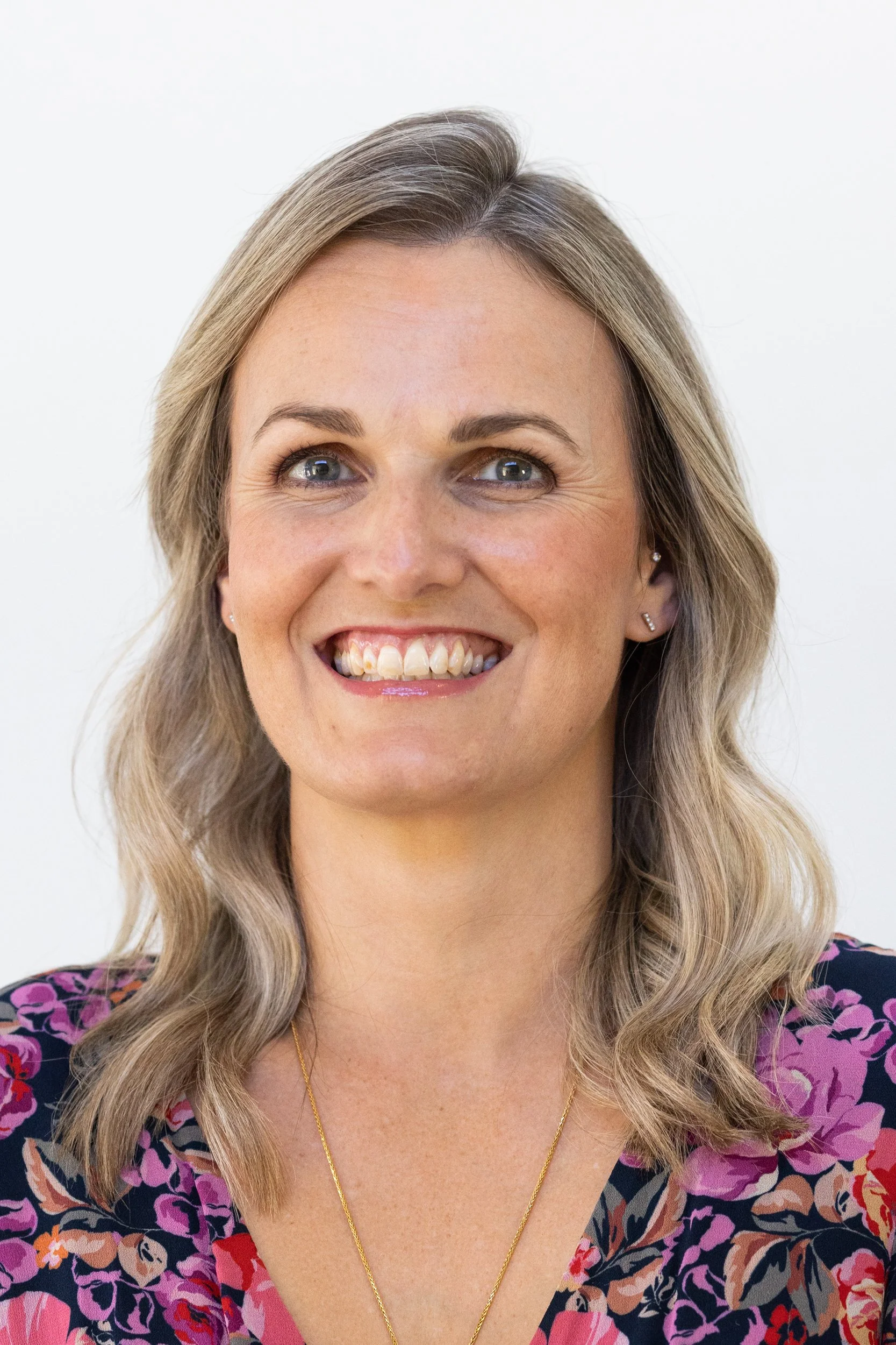 Close-up of a smiling woman with blonde wavy hair, wearing a floral shirt and a gold necklace, against a plain white background.