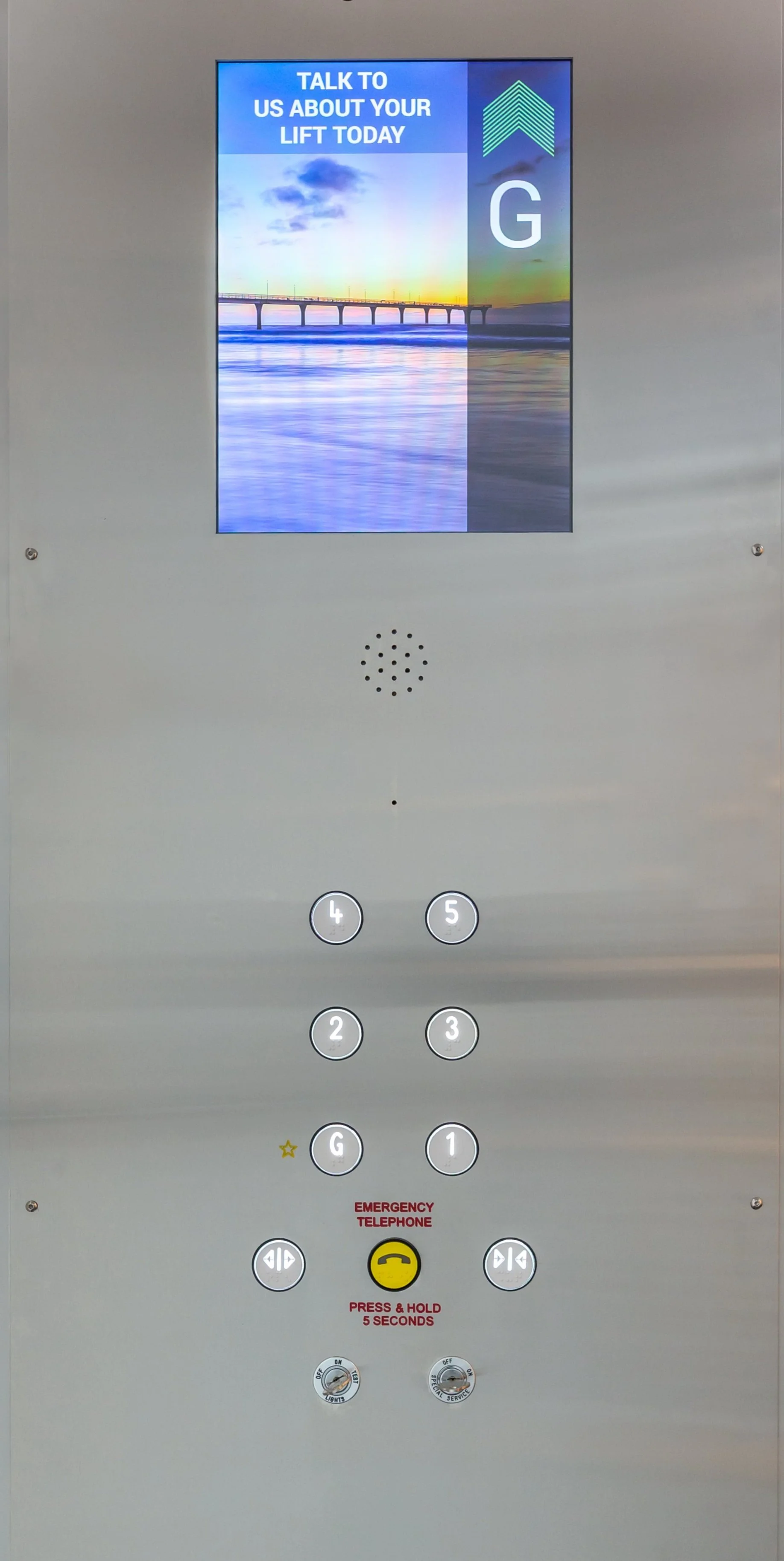 Elevator control panel with a digital screen displaying a message about talking to us about your lift today, buttons labeled 1, 2, 3, 4, 5, G for ground level, a yellow emergency telephone button, and the control switches for lighting and service.