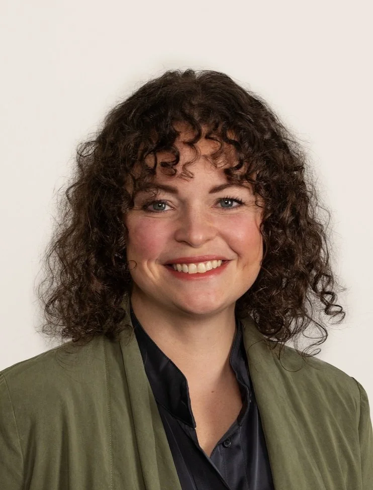 A woman with curly brown hair, blue eyes, and a friendly smile, wearing a dark shirt and an olive green jacket, standing against a white background.