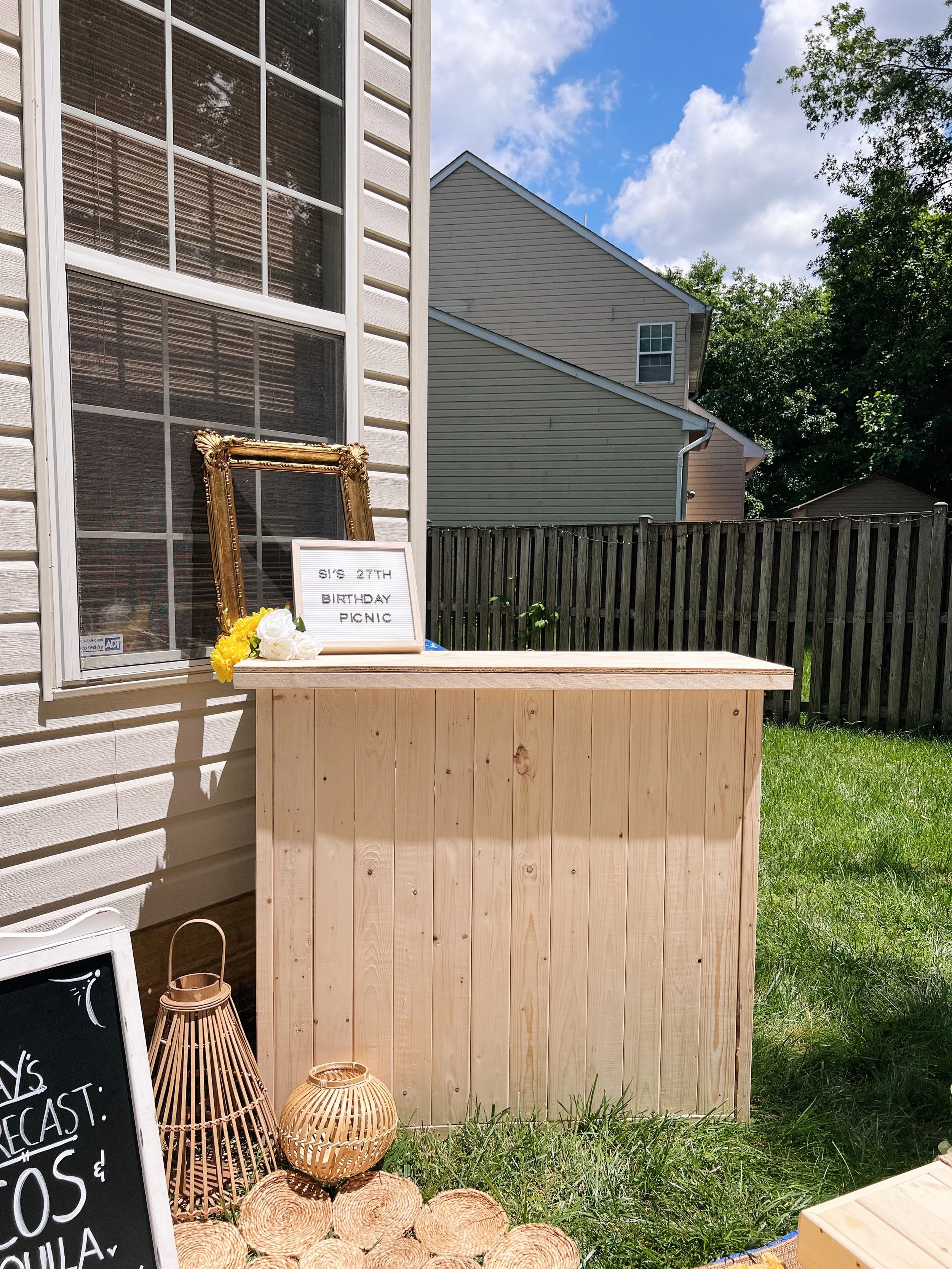 Pallet bar stand filled with assorted decorations, wines, candles, and dining sets on a picnic blanket with pillows designed by Picnics By Peace