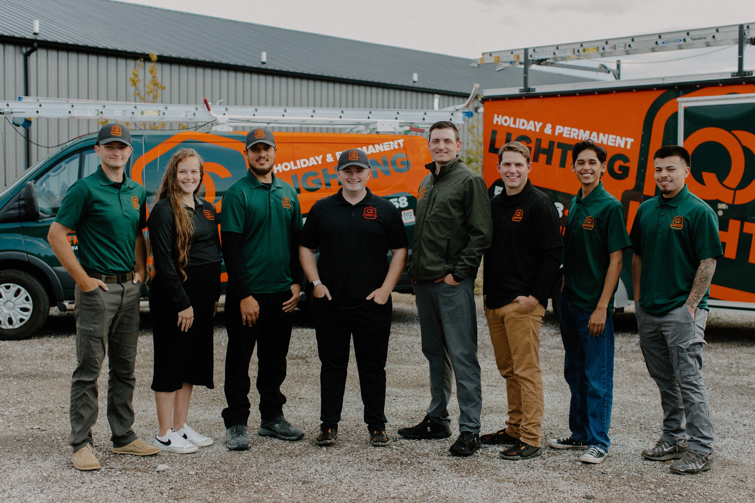 A group of nine people standing in front of a truck and van with orange and green branding for holiday and permanent lighting services. The group is smiling and wearing work uniforms, with some in green shirts and others in black, all with a logo.